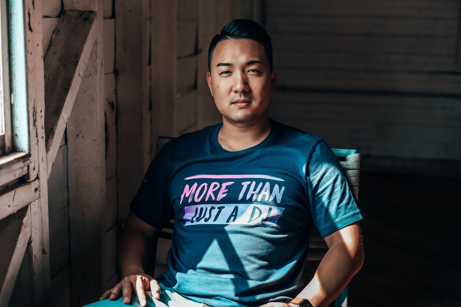 Indoor portrait of a man sitting in warm window light wearing a blue graphic tee, captured in a minimal, moody, lifestyle-inspired setting