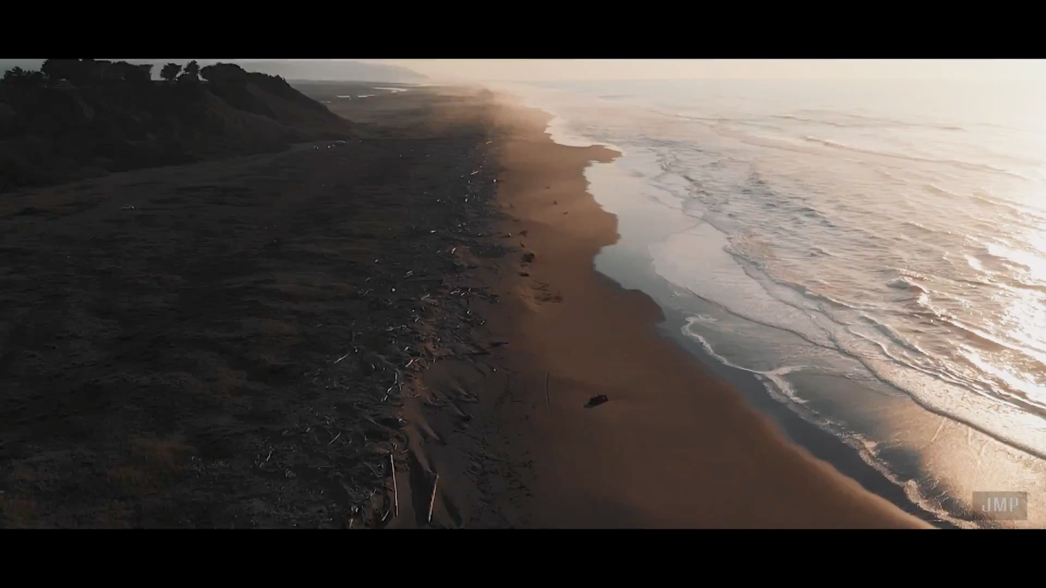 Aerial drone shot of a rugged coastline with dark sand and ocean waves at sunset, capturing dramatic light and sweeping shore.