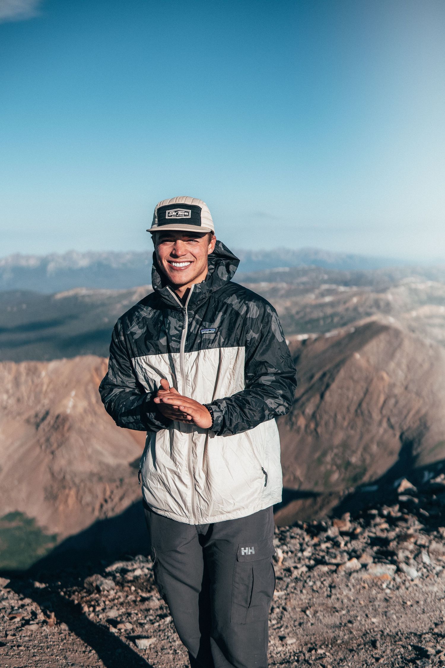 Adventure portrait of a smiling hiker on a mountain summit, surrounded by rugged peaks and bright morning light in a minimal outdoor style
