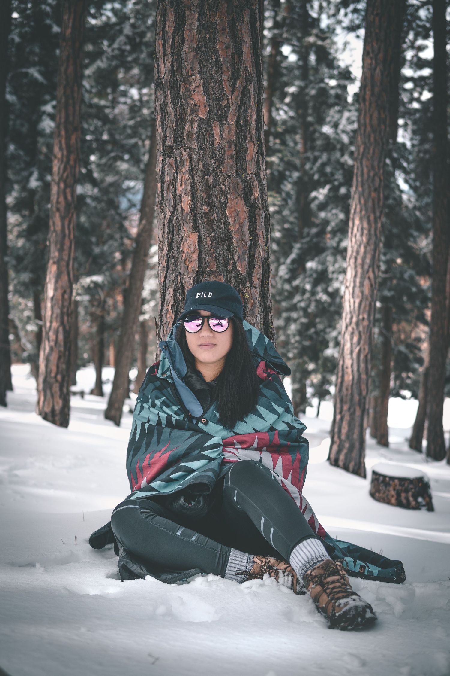 Woman sitting on a snowy forest pathway with tall pine trees and soft winter light creating a peaceful cold-weather adventure setting.