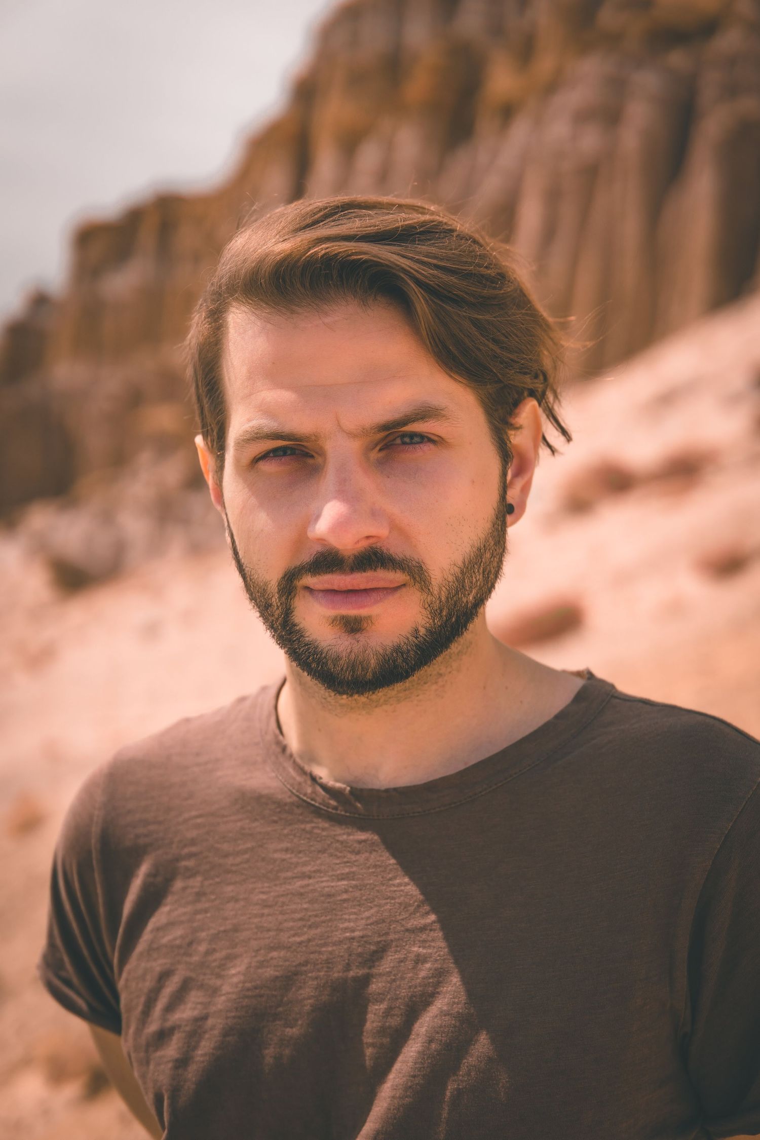 Man standing in a sunlit desert landscape with rugged cliffs behind him, giving a strong, natural portrait in warm earthy tones.
