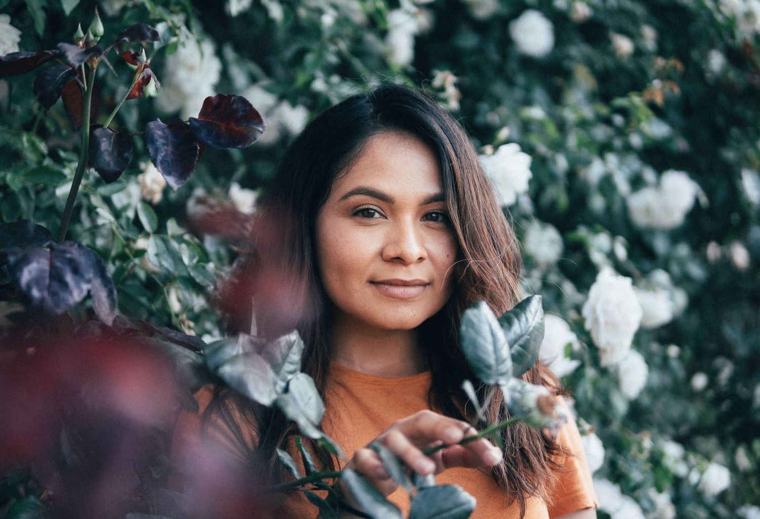 Natural outdoor portrait of a woman standing among blooming flowers, framed by greenery and soft bokeh for a serene botanical atmosphere.