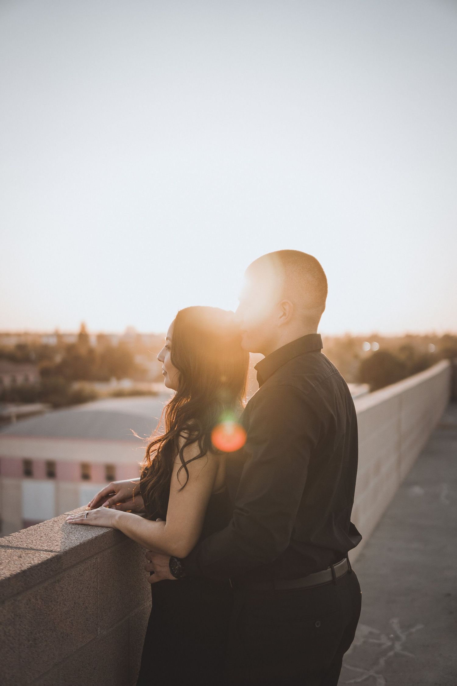 Romantic rooftop engagement photo of a couple embracing while watching the sunset, with warm golden sun flare highlighting their silhouettes