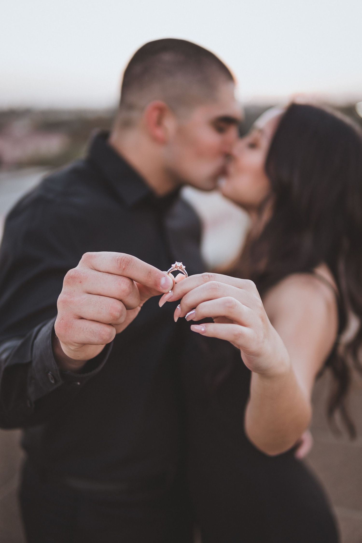 Close-up engagement photo of a couple kissing while holding a ring toward the camera, capturing romantic focus on the ring with soft sunset