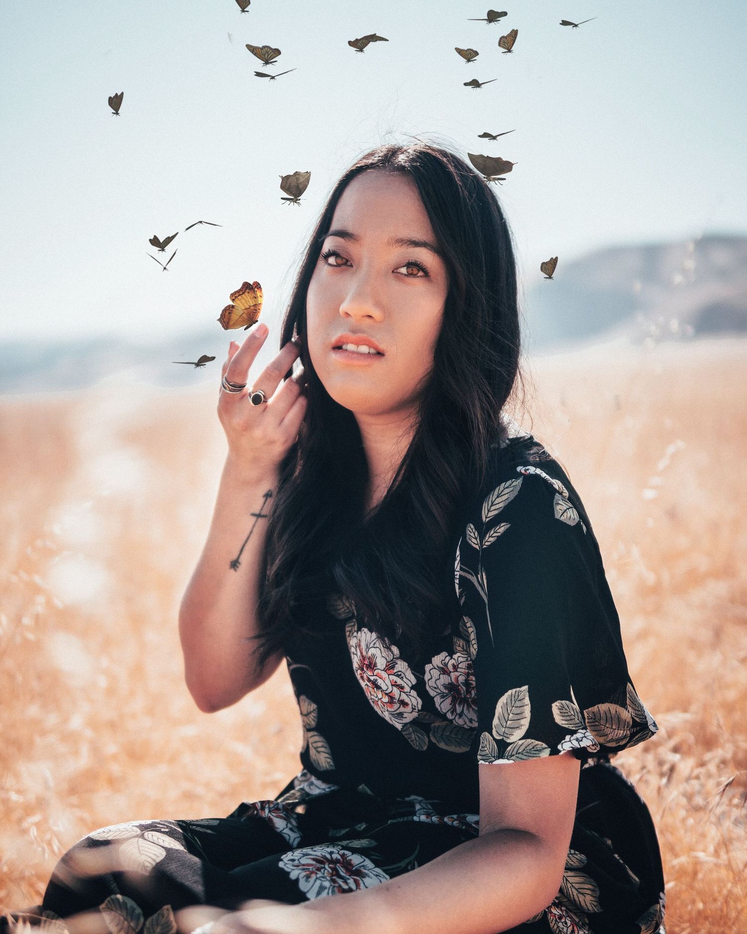 Woman sitting in a golden field as butterflies flutter above her, creating a dreamy, whimsical, nature-inspired portrait with soft light.