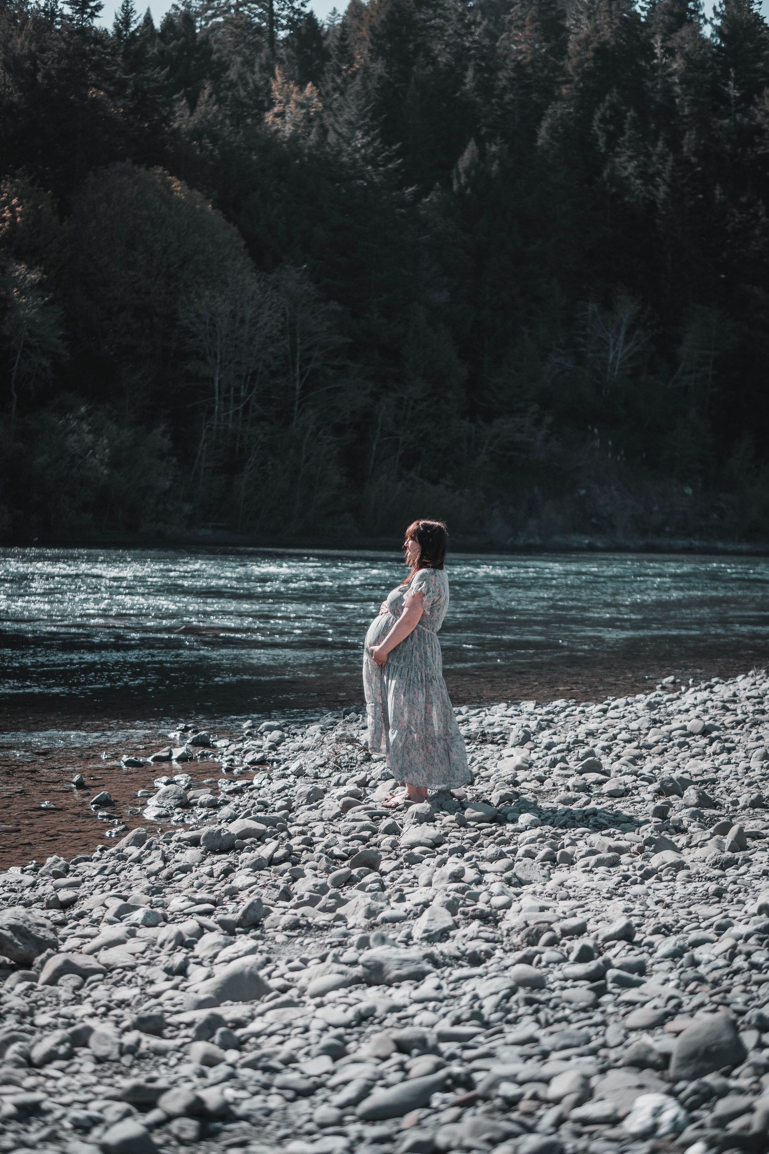Expectant mother in a flowing dress standing on a rocky riverbank with forested hills behind her, bathed in soft natural light.