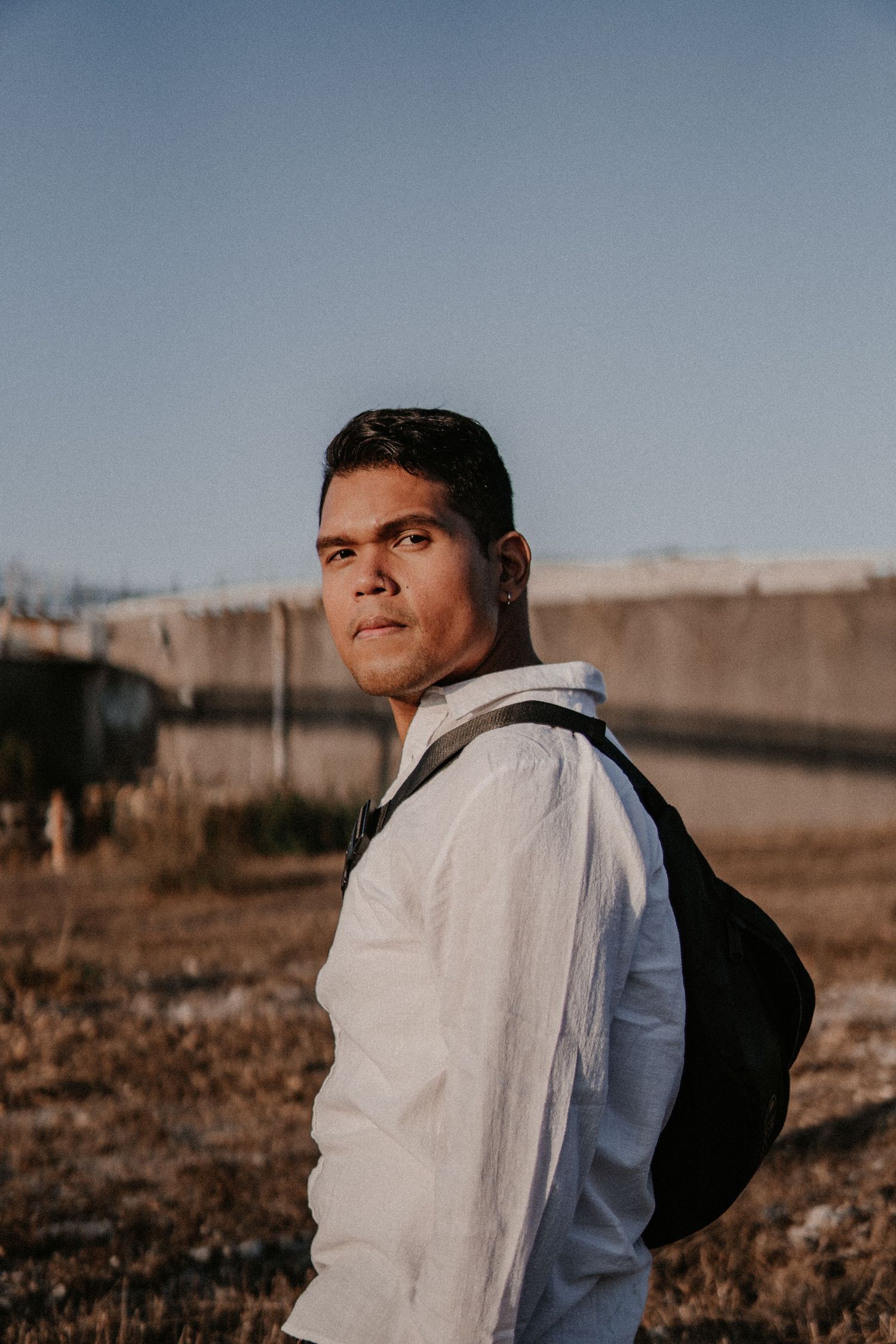 Man outdoors in golden-hour light wearing a white shirt and backpack, looking over shoulder against a blue sky.