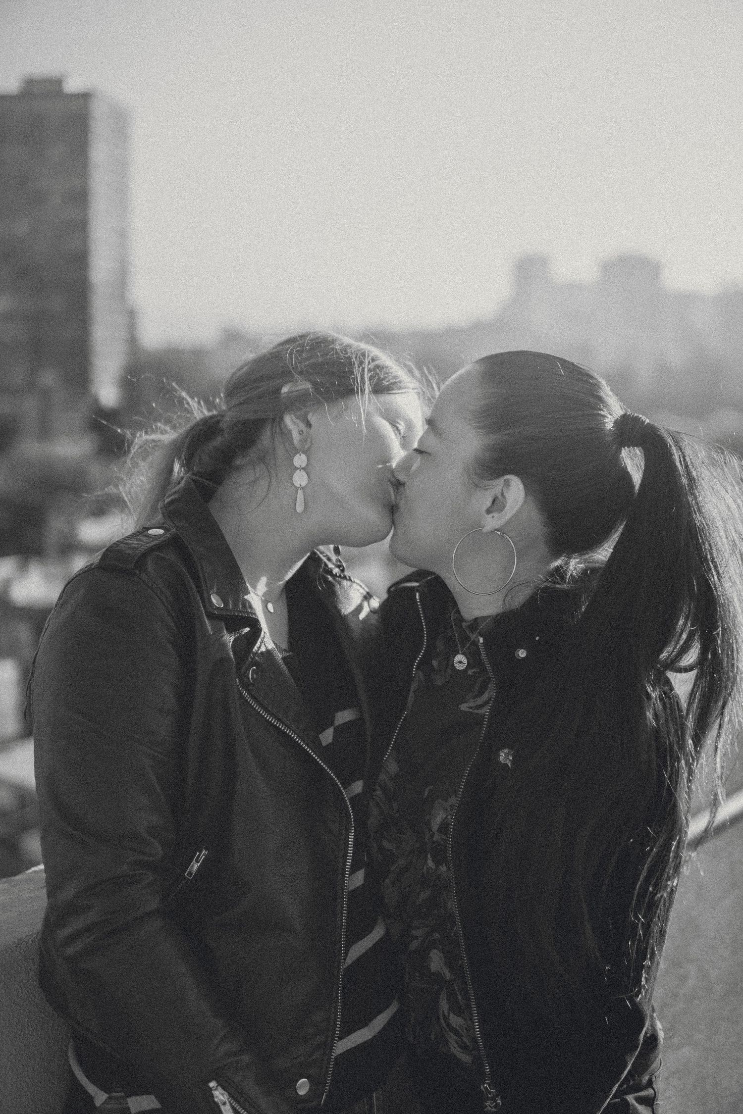 Romantic black-and-white photo of a couple sharing a kiss on a rooftop with a soft, blurred city skyline glowing behind them