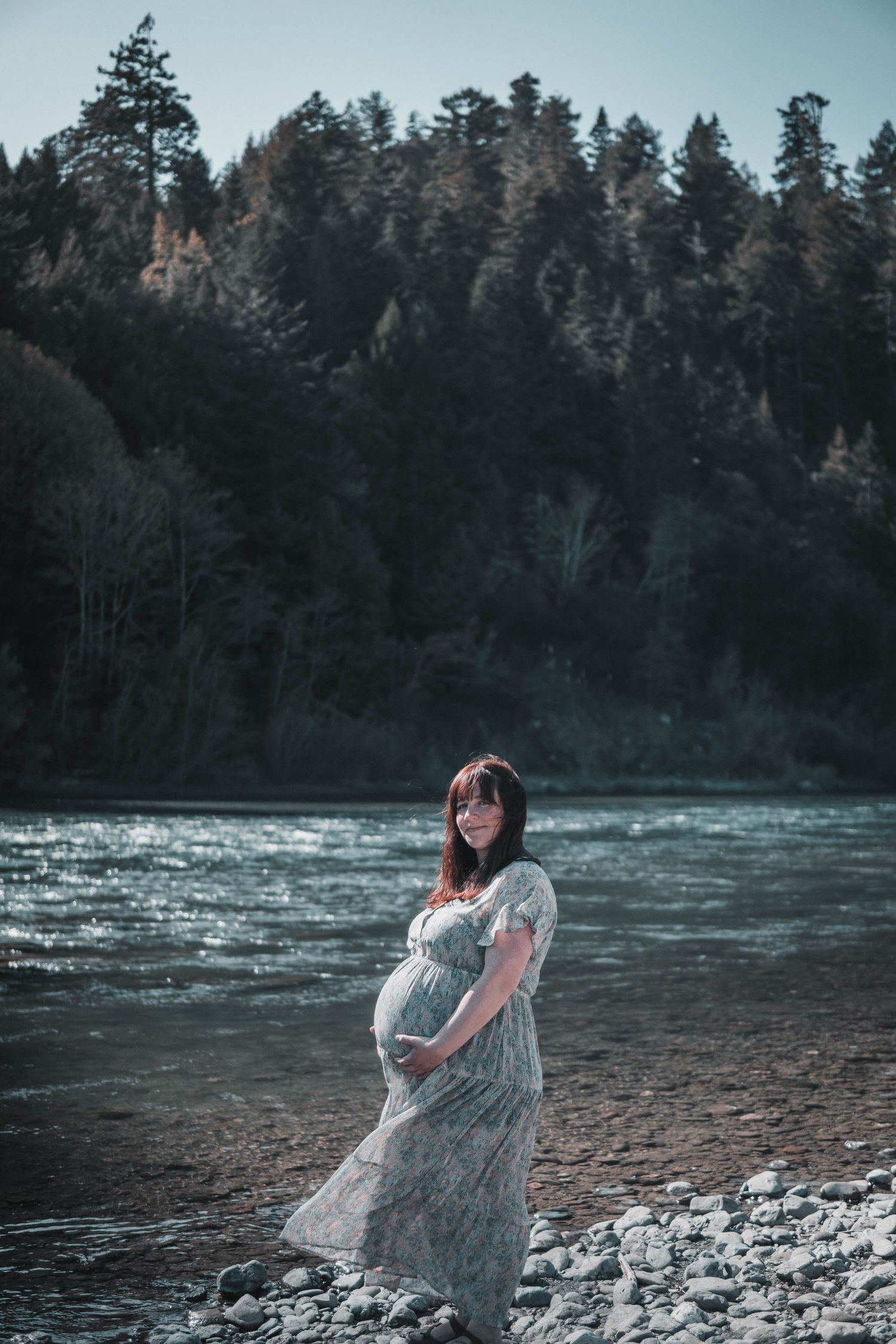 Pregnant woman in a floral dress holding her belly along a rocky riverbank, gazing at shimmering water beneath tall evergreen trees
