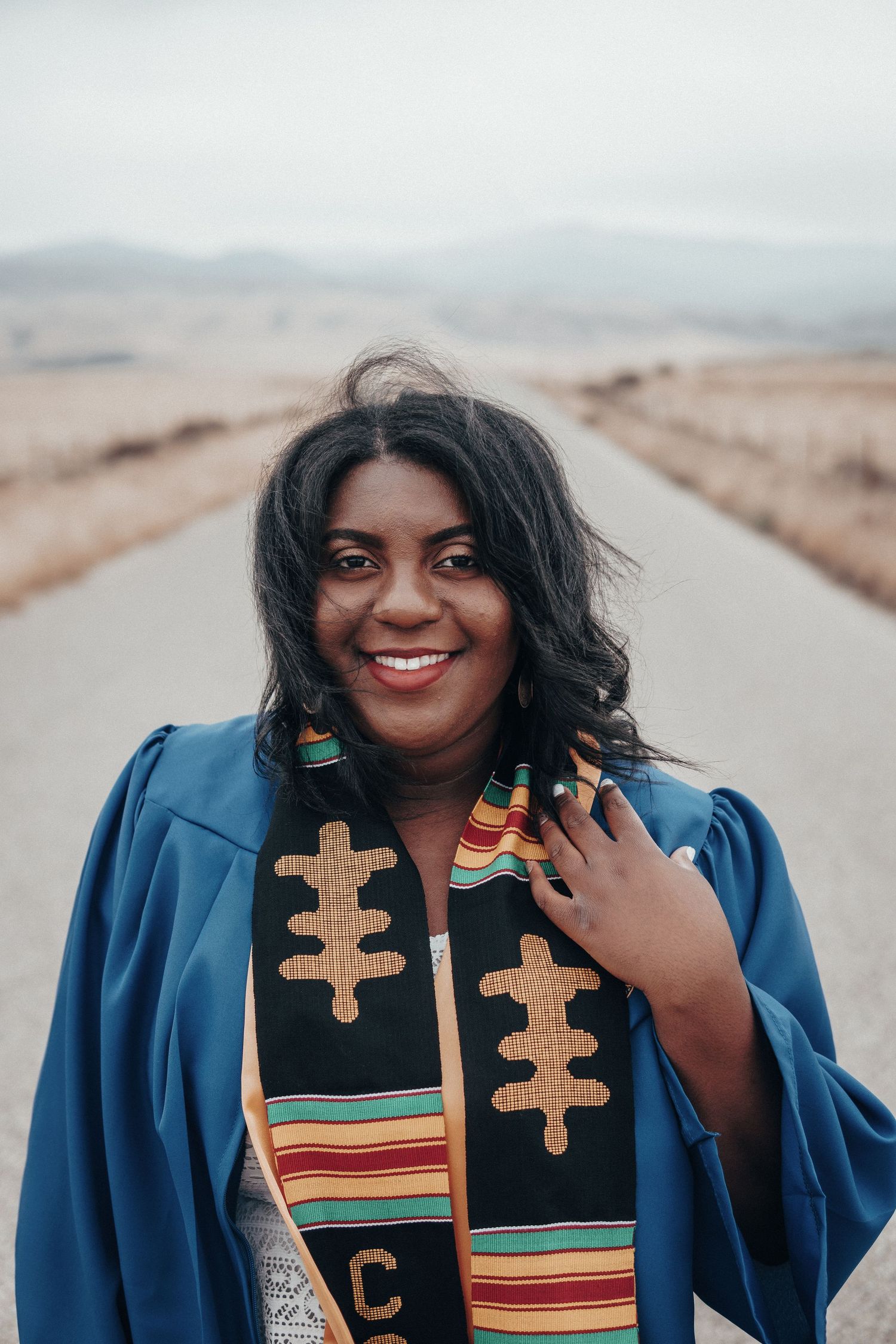 Smiling graduate in a blue gown and colorful kente stole standing on a rural road with soft mountains behind her, celebrating achievement.