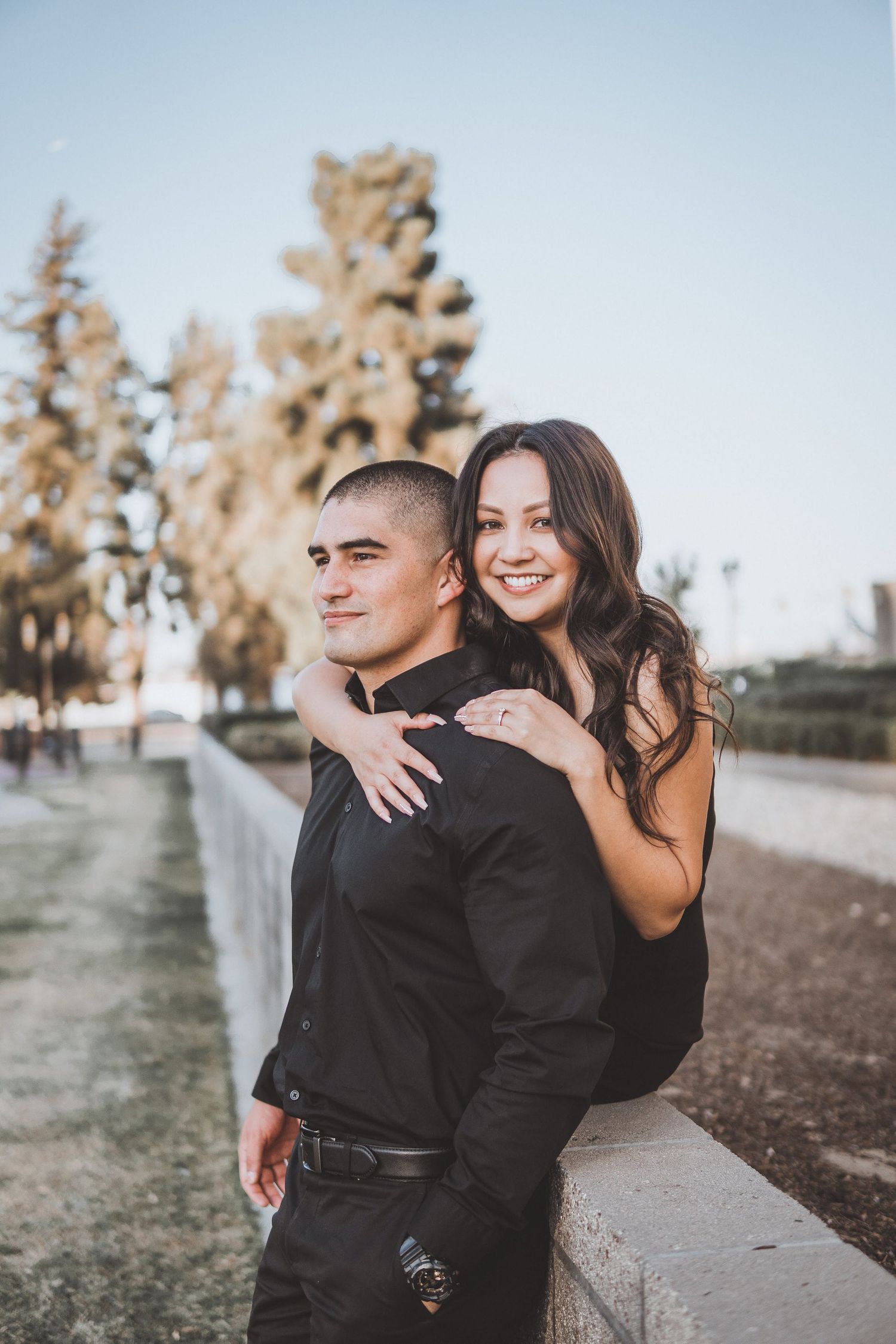 Engagement photo of a smiling couple in black outfits, with the woman hugging her partner from behind along a walkway lined with trees.