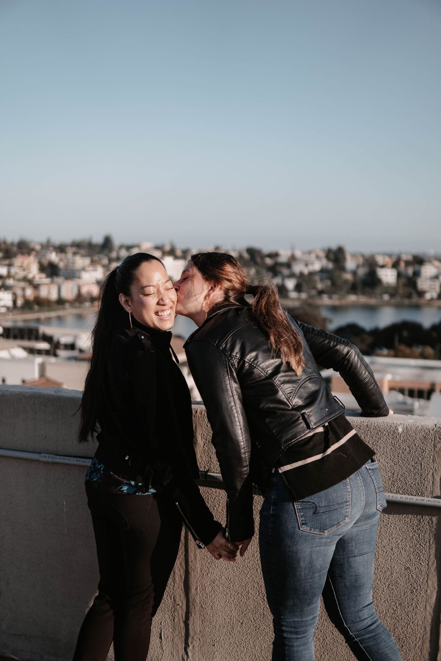 Playful moment of a couple holding hands as one leans in for a kiss on a bright rooftop overlooking a sunlit city and water.