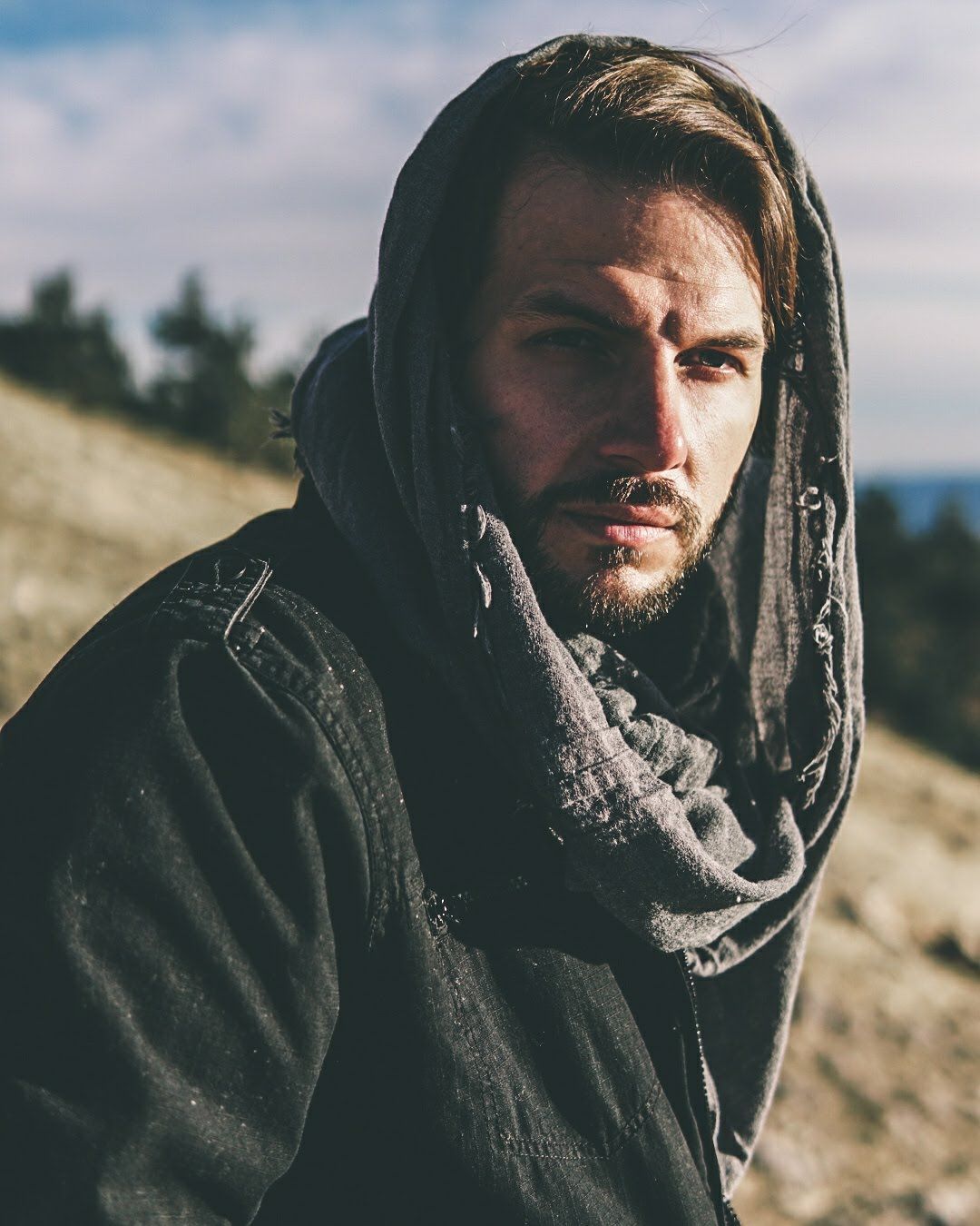 Man wearing a hooded scarf and dark jacket standing in harsh sunlight on a rugged hillside, looking into the distance.