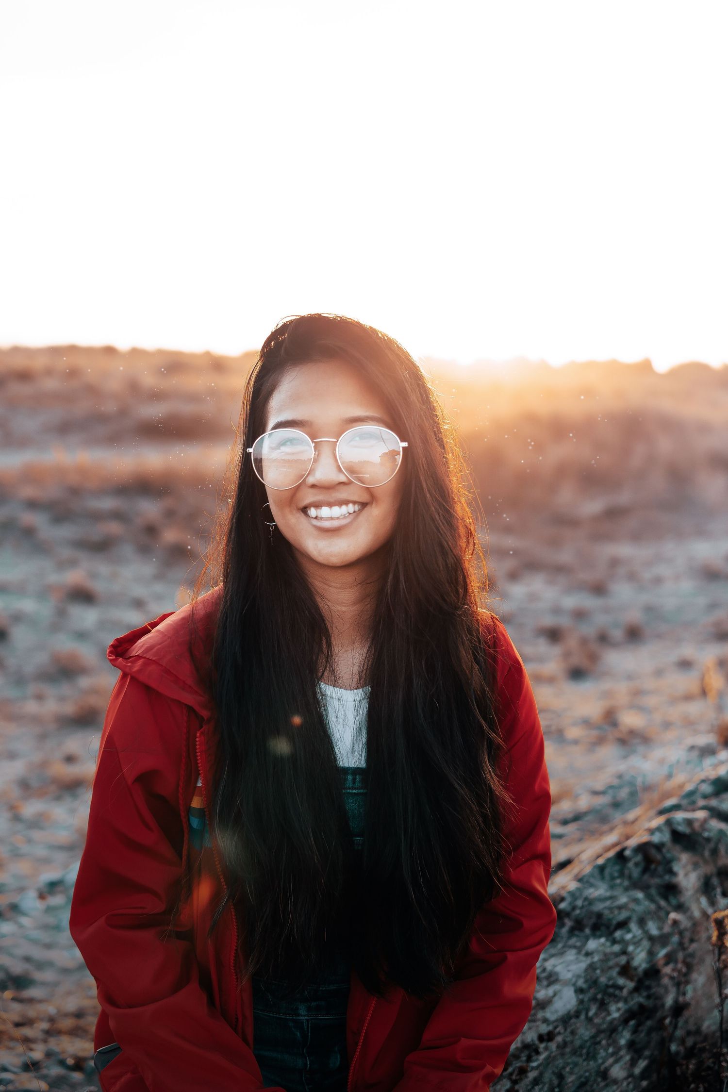 Minimal adventure portrait of a smiling woman in a red jacket at golden hour, embracing nature with warm light and serene outdoor vibes.