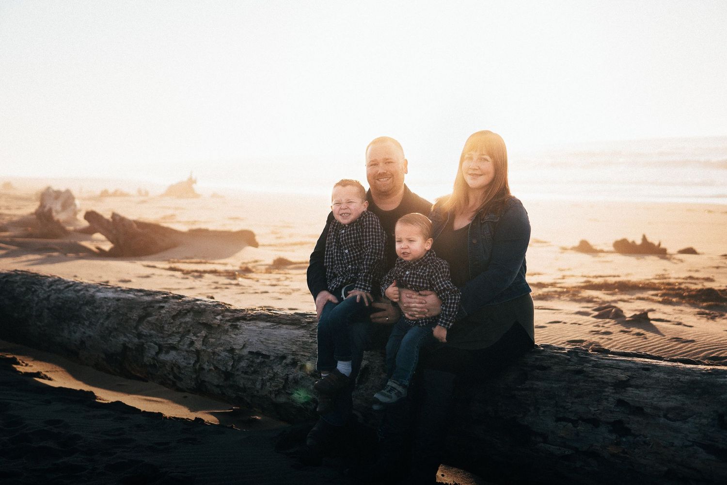 Family portrait at the beach during a warm sunset, with parents and two young children sitting on driftwood near the shoreline.