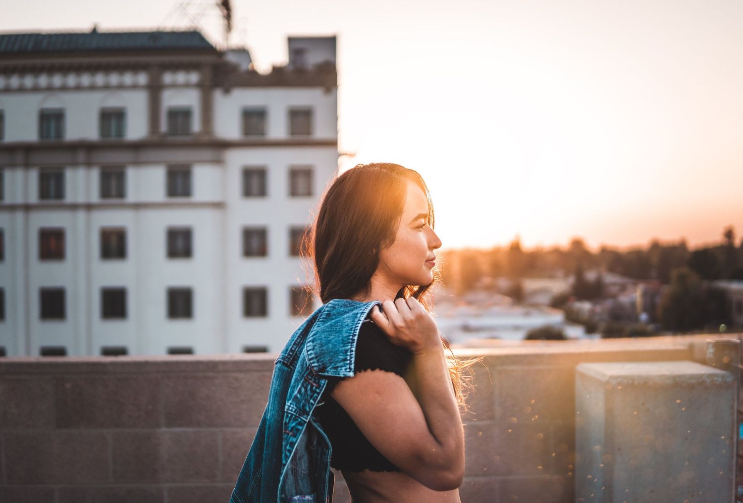 Woman standing on a rooftop at sunset with warm golden light behind her, holding a denim jacket and looking out over the city .