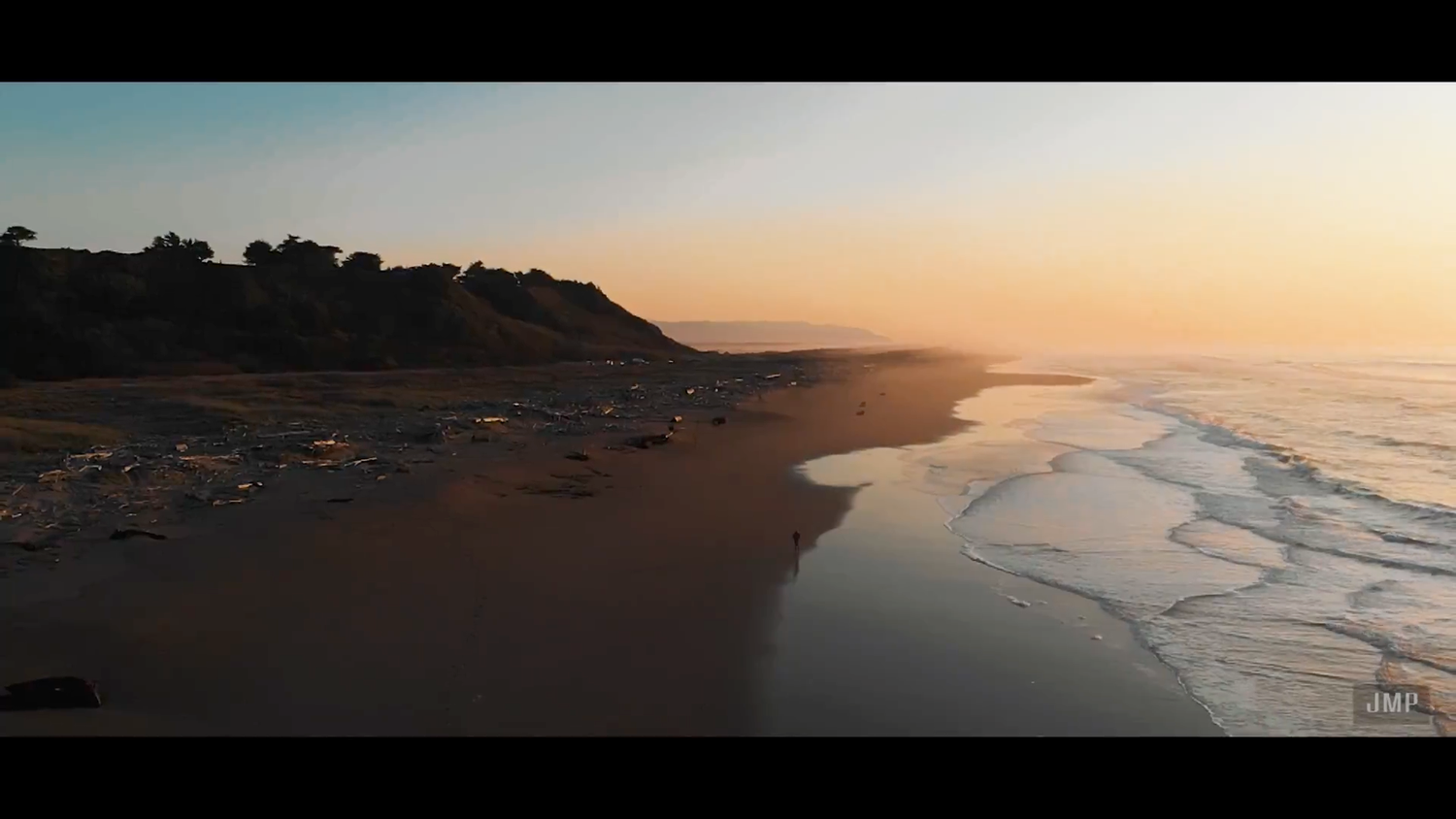 Golden hour drone view of a wide beach and calm shoreline with warm sunlight casting long shadows on the coastal landscape