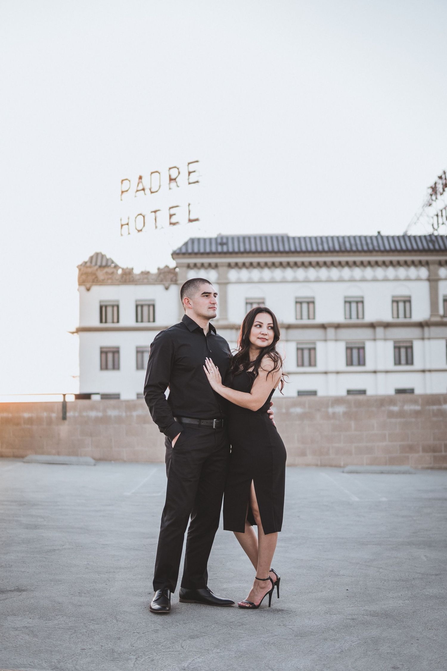 Elegant couple dressed in black posing on a rooftop with historic hotel backdrop, soft evening light creating a timeless romantic scene