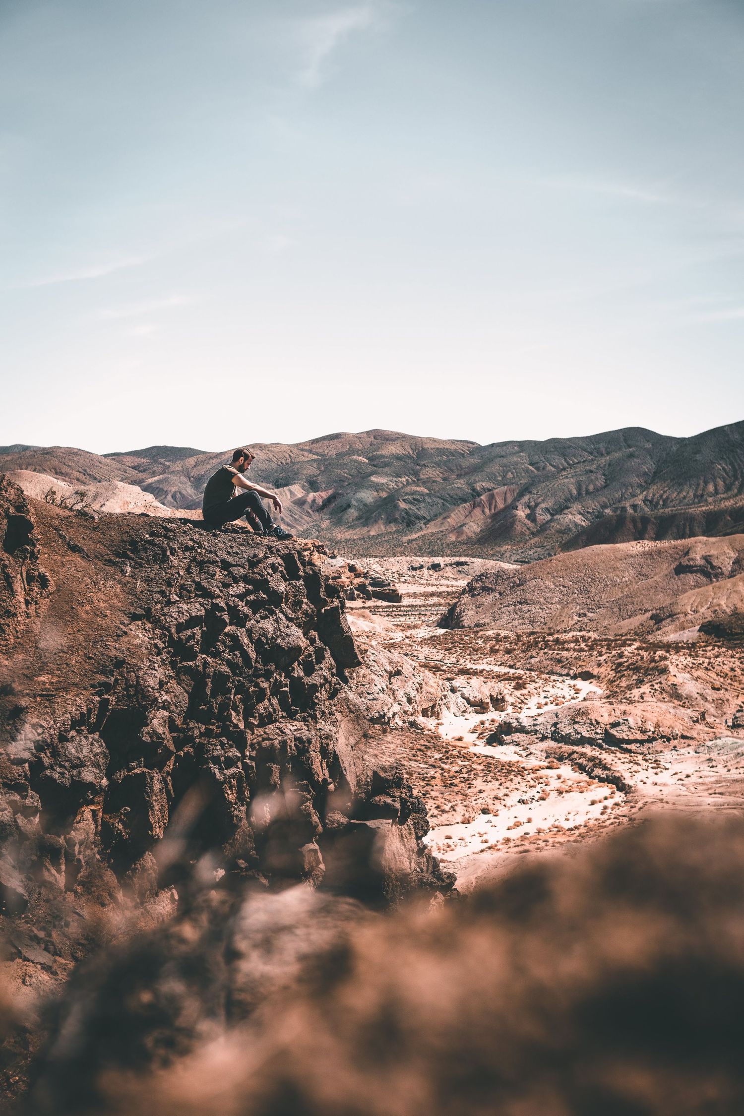 Person sitting on a rugged cliff overlooking vast desert mountains under a clear sky, capturing a peaceful outdoor adventure and scenic wild