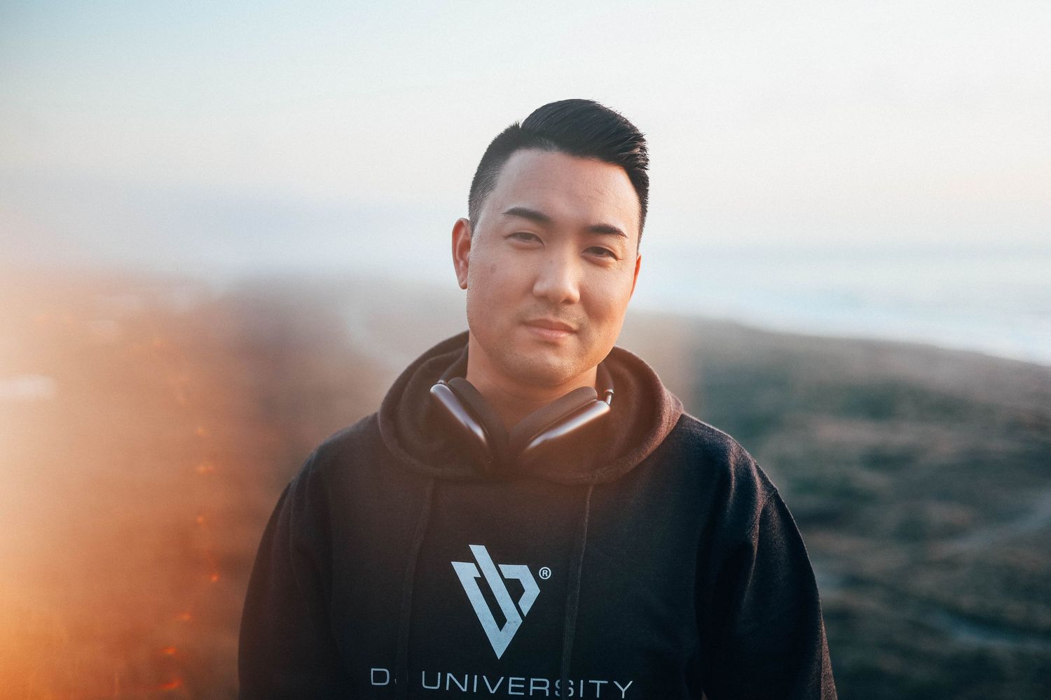 Outdoor portrait of a man in a black hoodie with headphones resting on his neck, standing near the coast at sunrise with warm soft light.