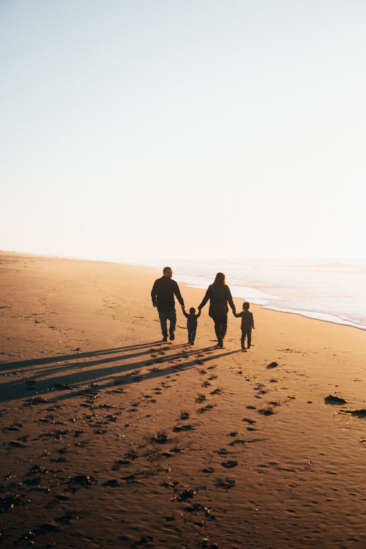 Family walking hand in hand along the beach at sunset, creating long shadows on the sand with soft ocean light and peaceful coastal vibes