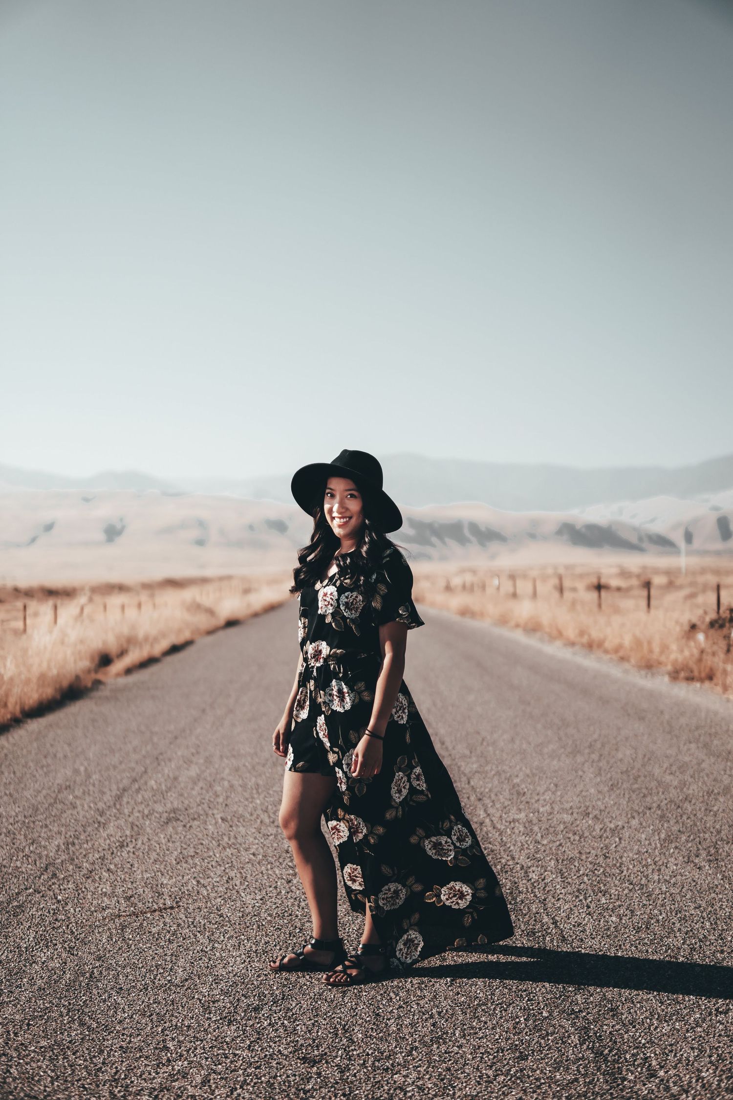 Woman in a floral dress and hat standing on an empty desert road with mountains in the distance, creating a stylish outdoor travel portrait