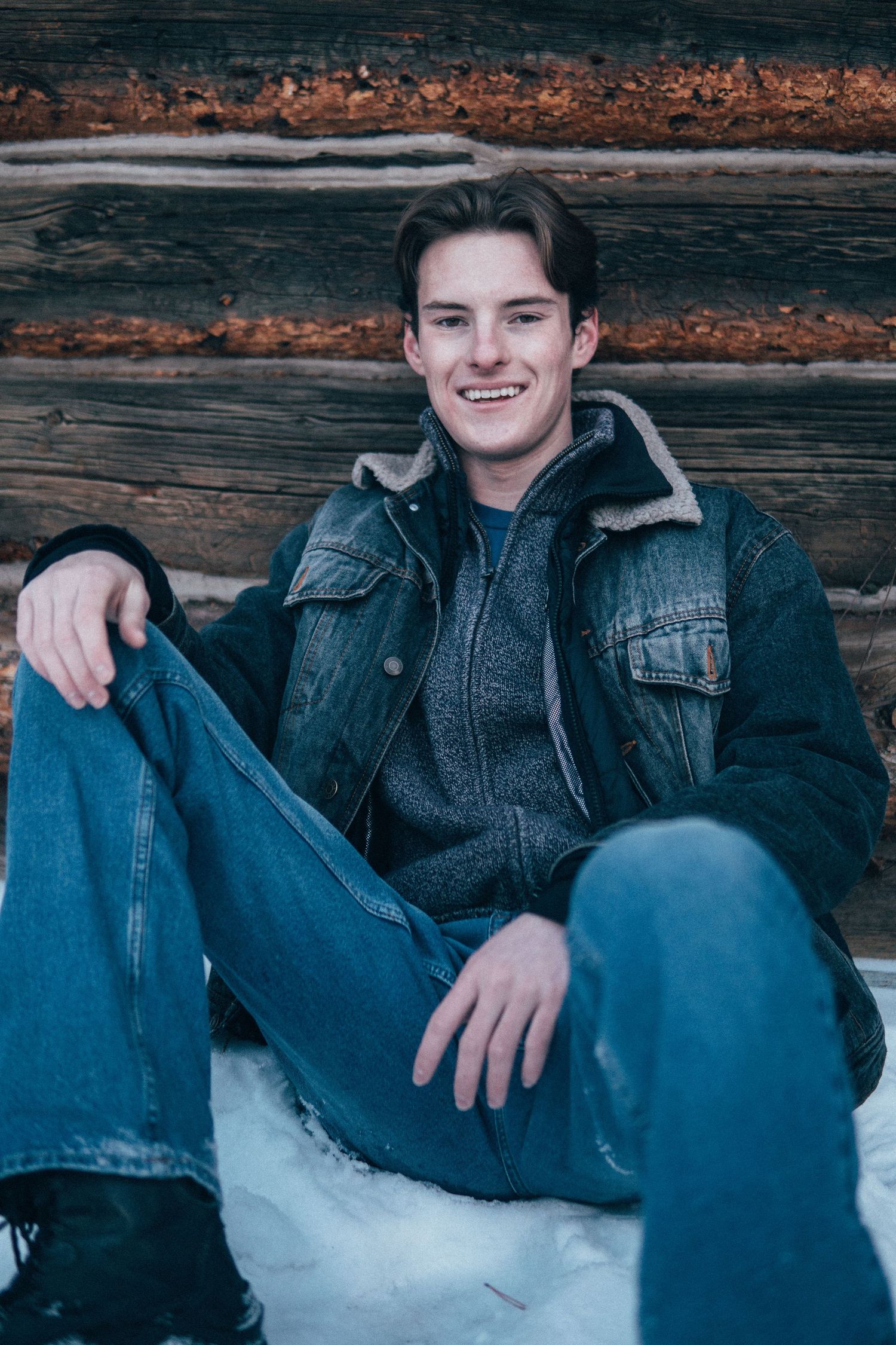 Man sitting against a rustic wooden cabin wall on winter snow, wearing layered denim and cold-weather clothing in a cozy outdoor setting