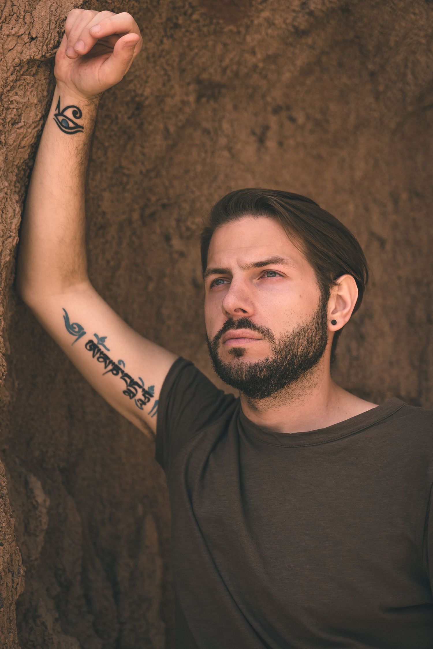 Man with short hair and arm tattoos posing against a textured rock wall delivering earthy, edgy, minimalistic atmosphere.