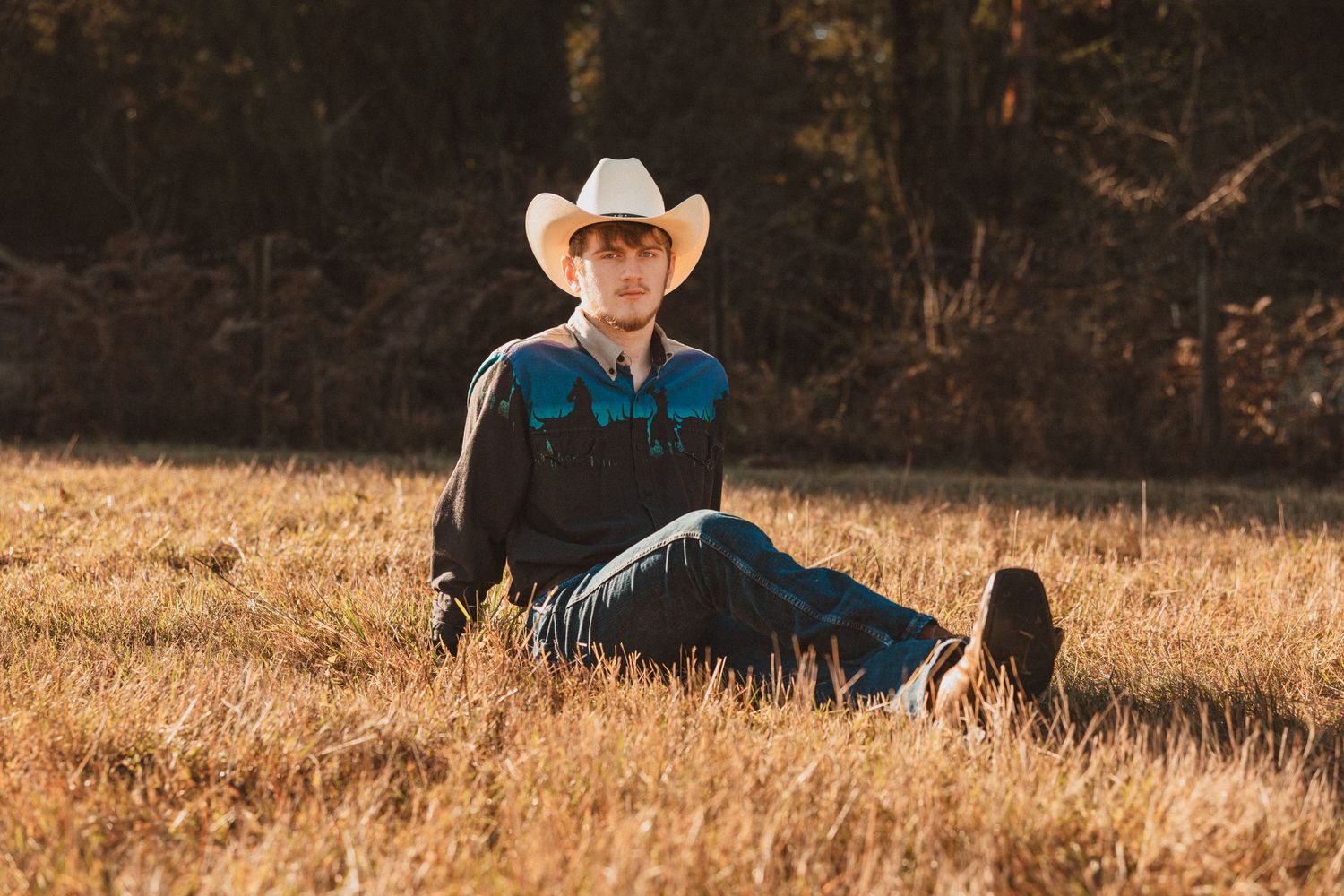 A person in western attire with a cowboy hat relaxes in a golden field during sunset.