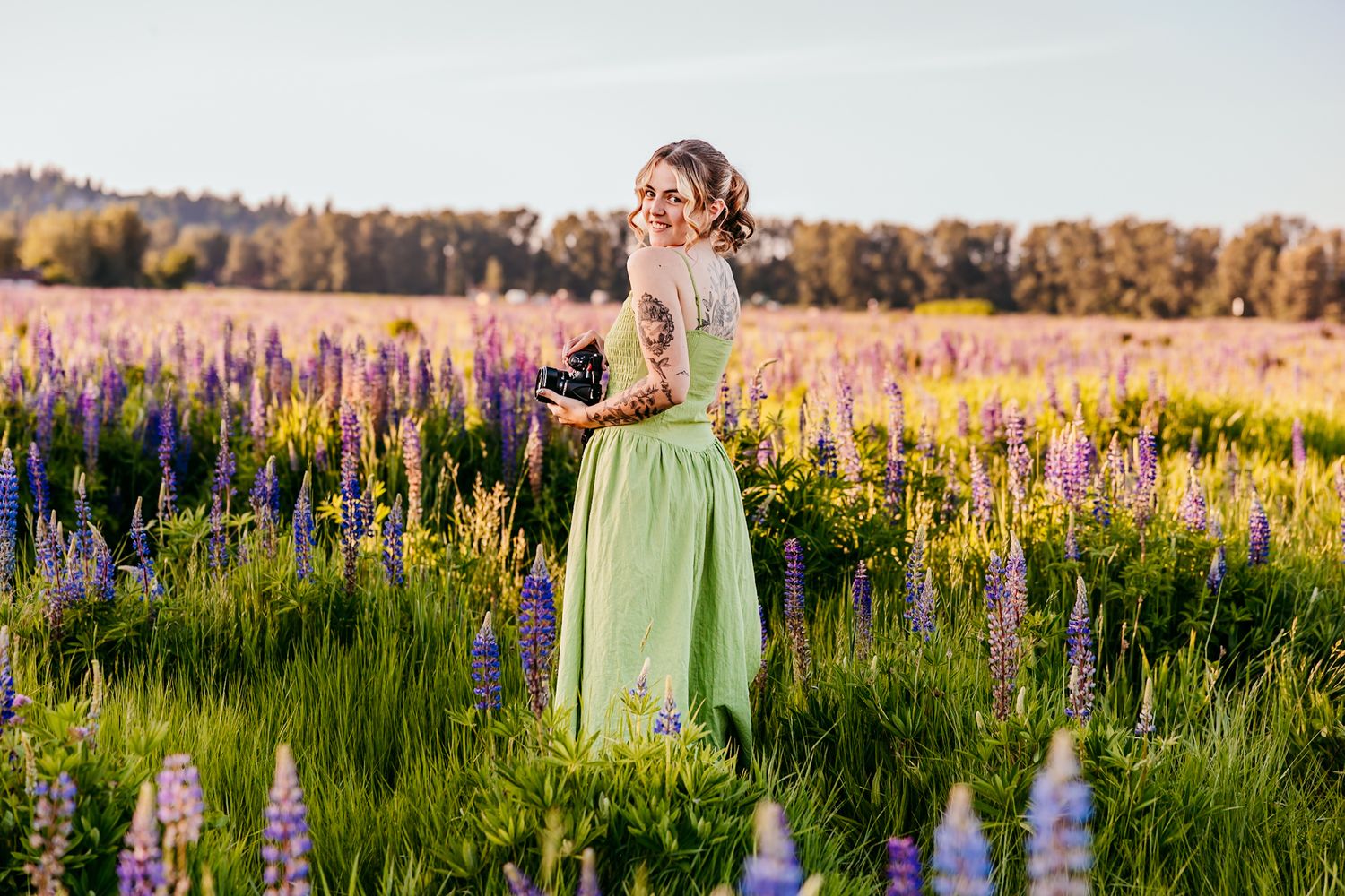 Woman in green dress standing in vibrant purple lupine flower field during golden hour sunset.