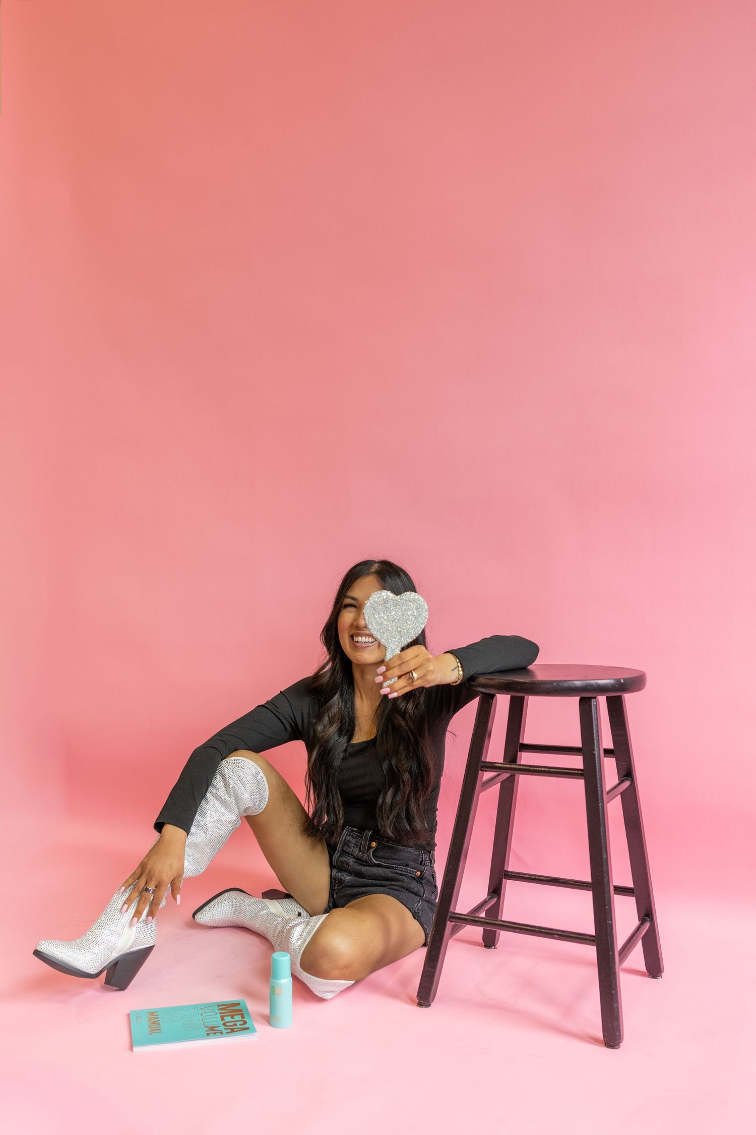 Stylish figure in black outfit and white boots poses on floor next to wooden stool against pink studio backdrop.