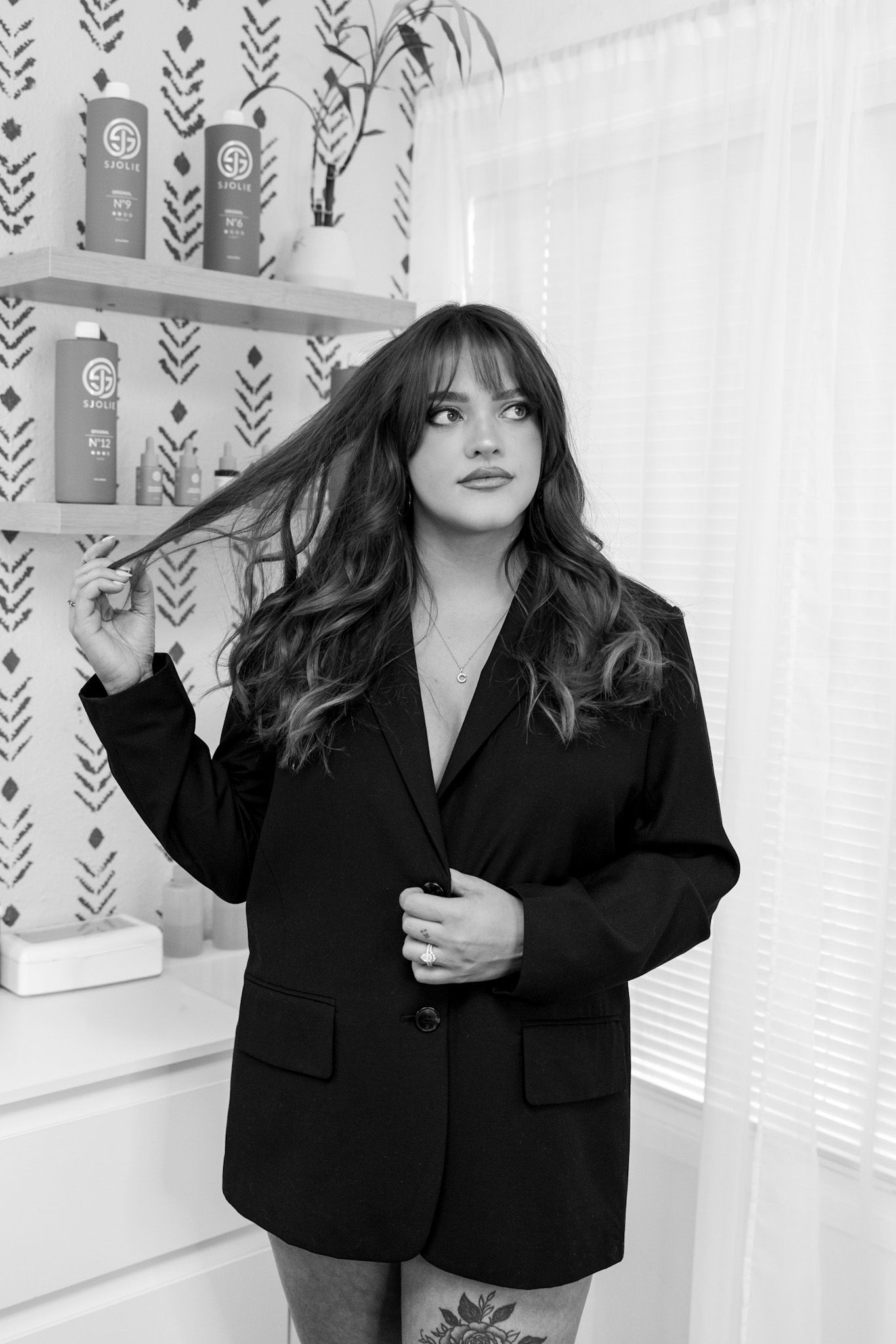 Black and white photo of someone in an oversized blazer posing against a wall with decorative wallpaper and shelving.