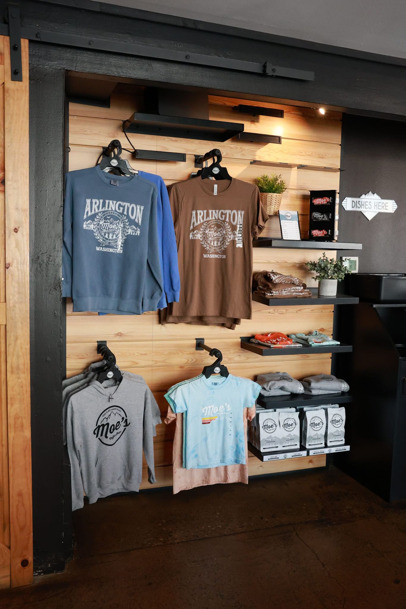 Retail display wall with Arlington branded sweatshirts and Moe's t-shirts on wooden shelving in a store.