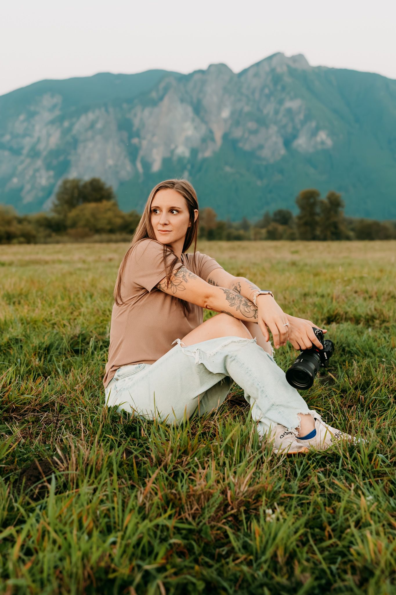 Woman with tattoos sits in green field holding camera, with dramatic mountain backdrop at golden hour.