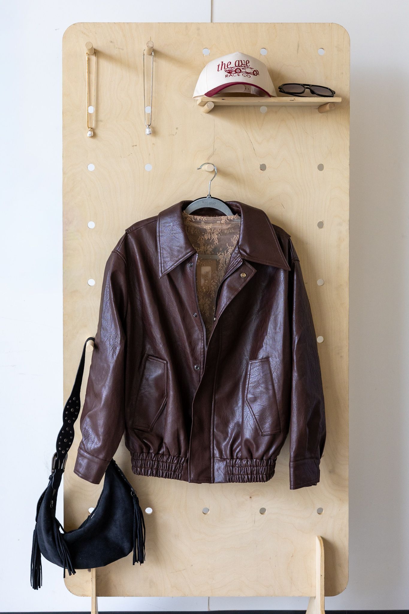 Brown leather bomber jacket on hanger with black fringe bag, baseball cap, and sunglasses on wooden wall rack.