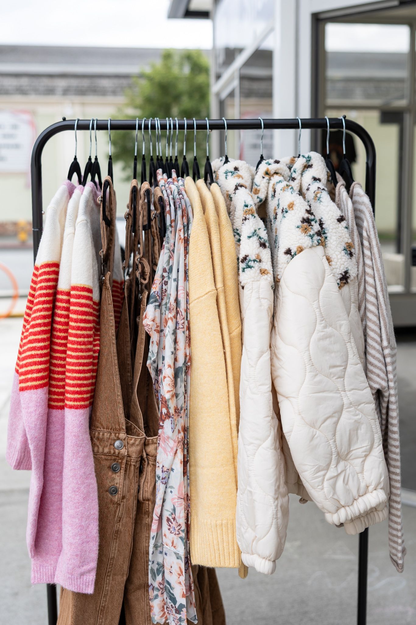 Outdoor clothing rack displaying colorful sweaters, floral blouses, and a white quilted vest on black hangers.