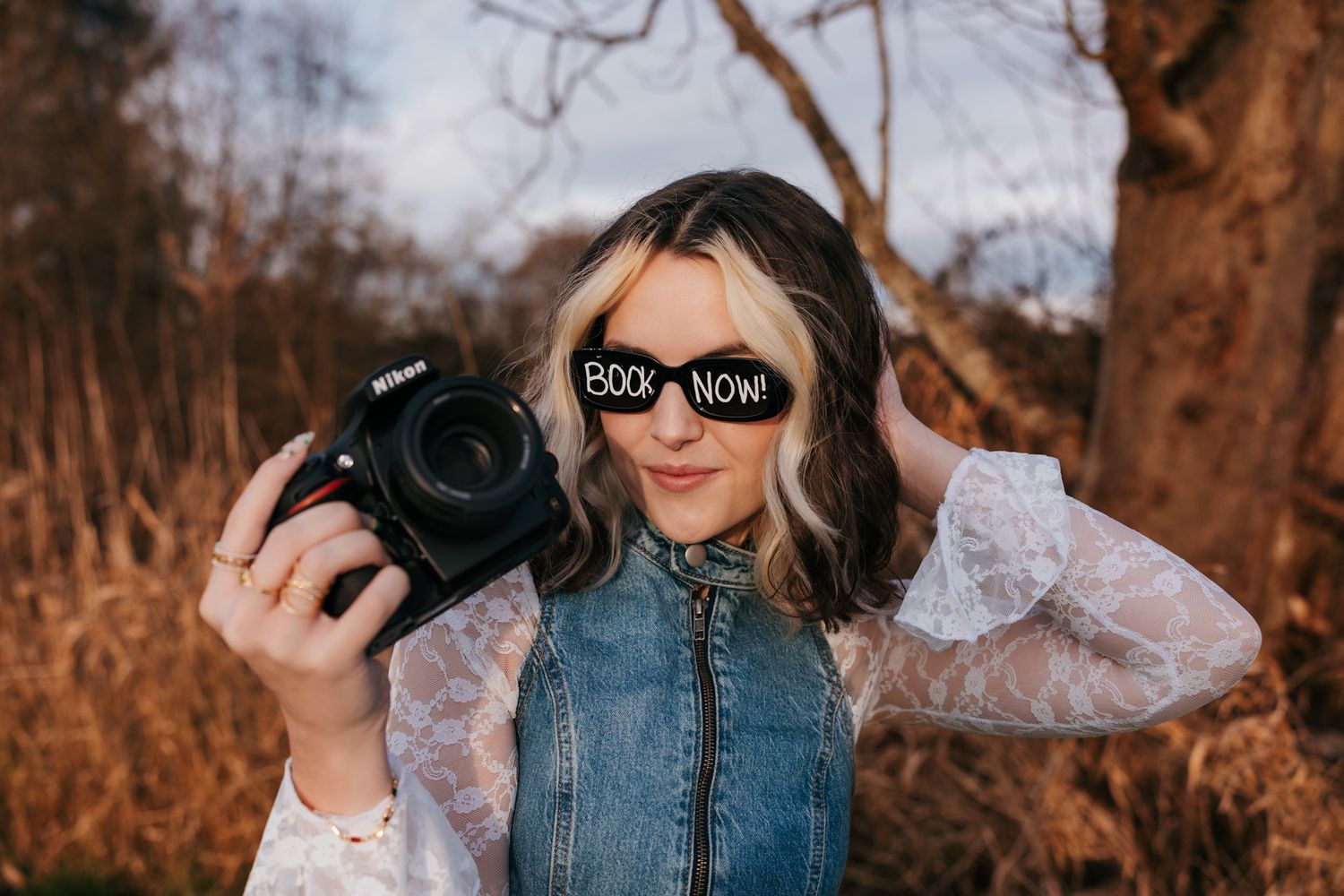 Person in denim jacket and sunglasses poses outdoors against autumn trees in warm golden light.
