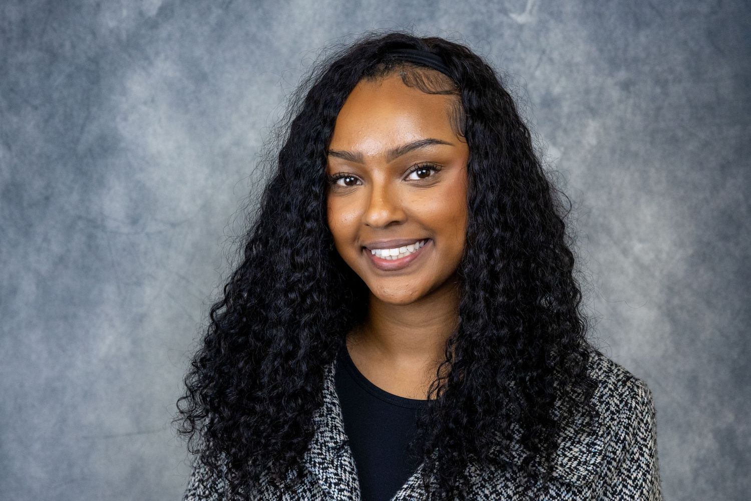 Professional headshot of a person with long curly black hair wearing a gray patterned blazer against a mottled background.