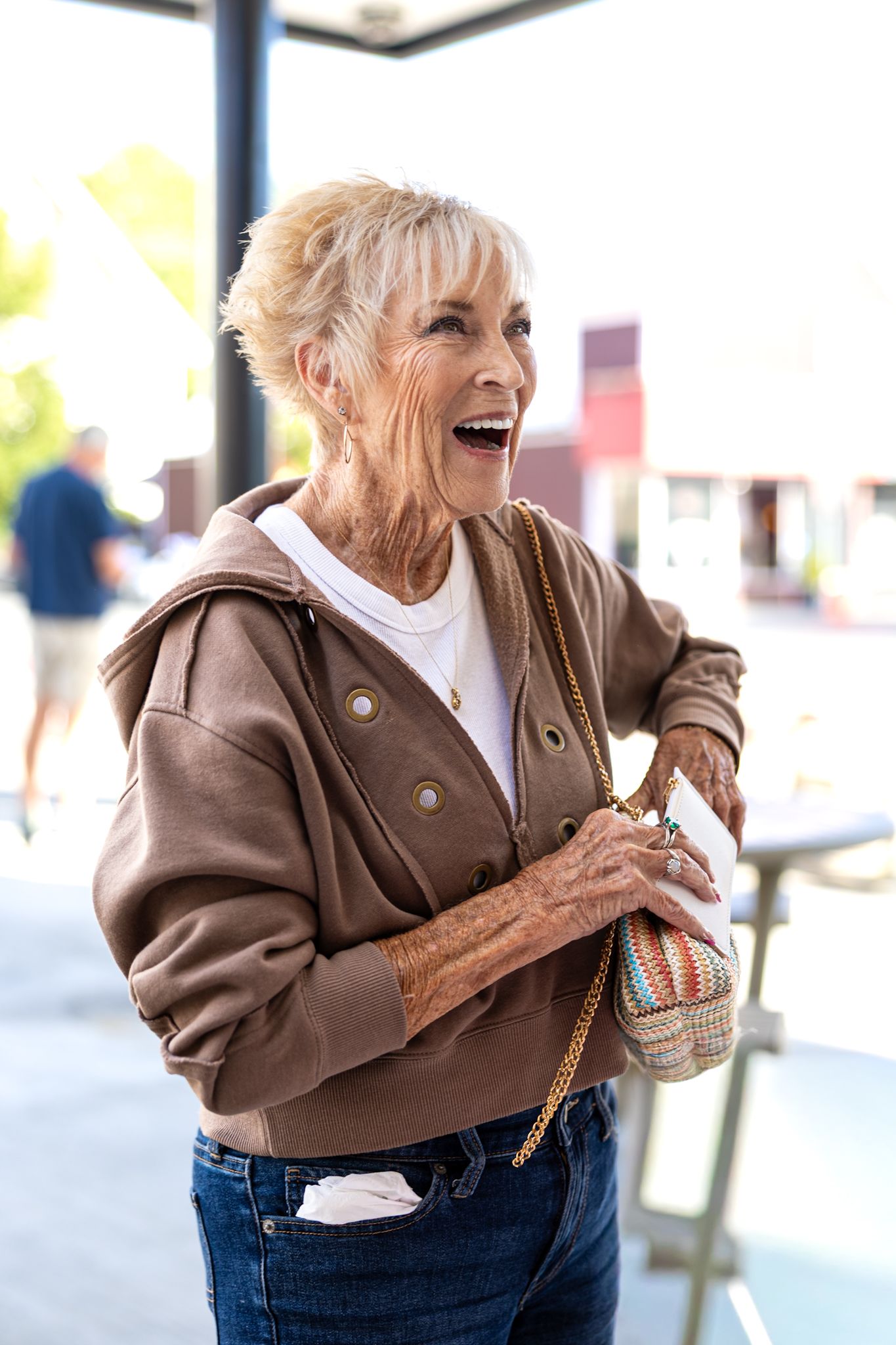 Cheerful senior woman in brown jacket laughing outdoors, holding colorful woven purse and wearing jeans.