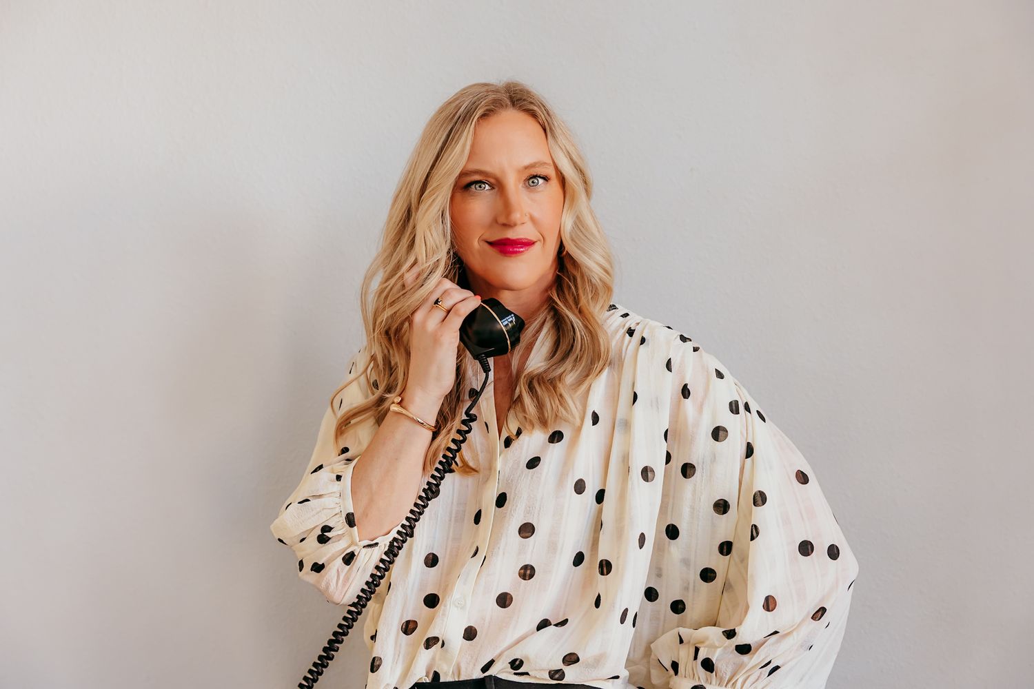 Blonde woman in polka dot blouse with red lips holding a vintage black telephone receiver.