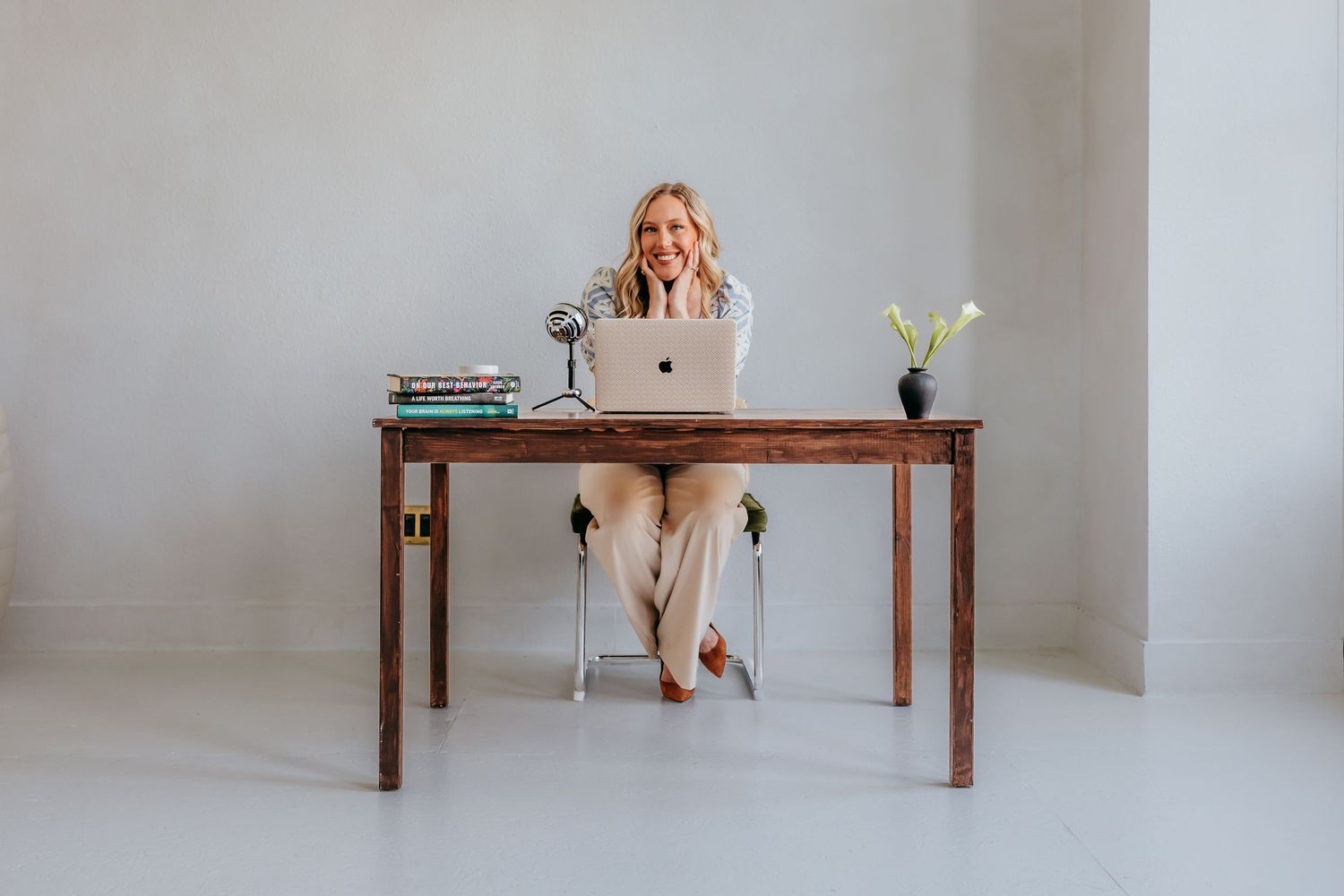 A smiling blonde woman sits at a wooden desk with a laptop, books, and a vase in a bright minimalist office.