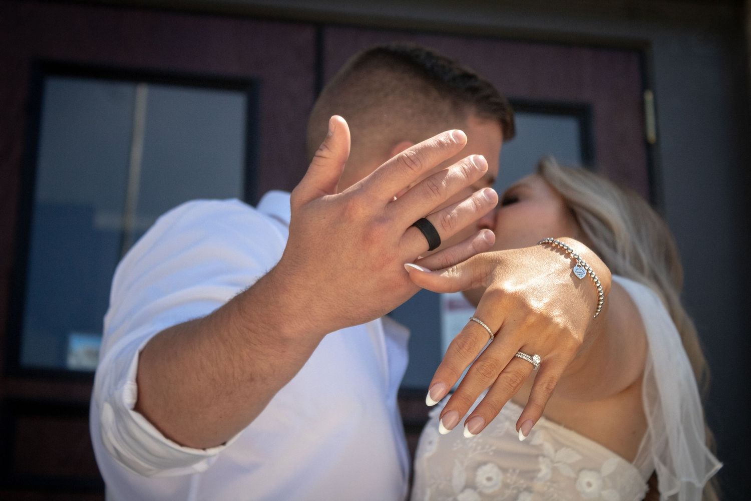 A couple wearing wedding attire show off their rings while playfully covering their faces with their hands.
