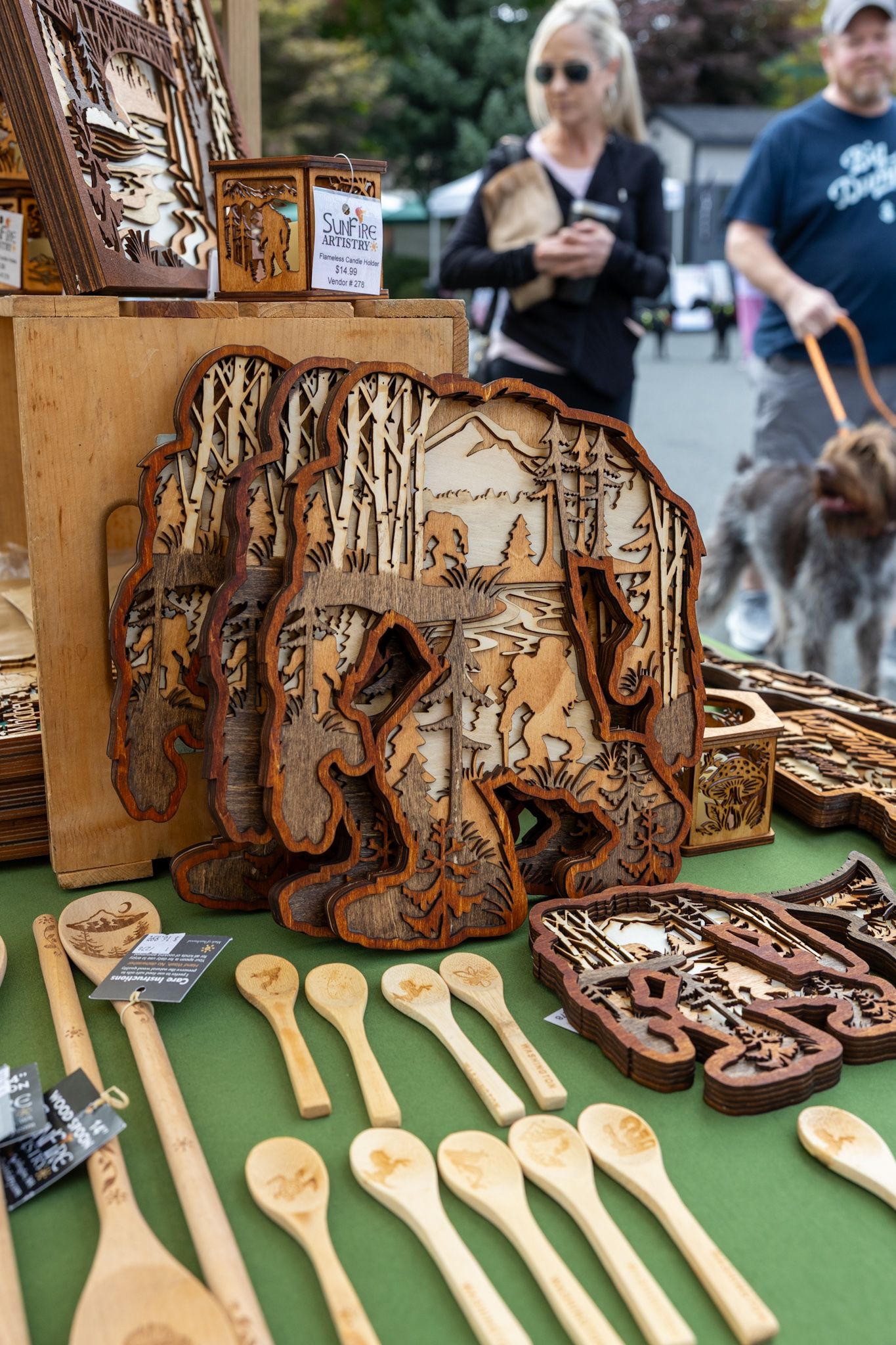 Handcrafted wooden Bigfoot laser-cut art pieces and engraved spoons displayed at an outdoor market vendor booth.