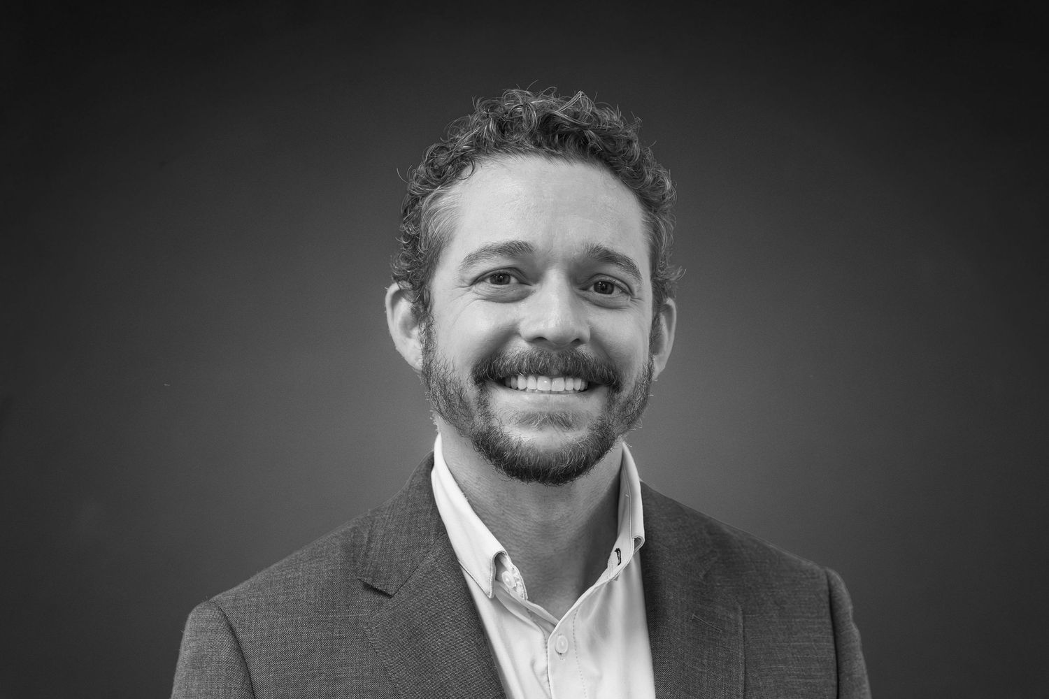 Professional black and white headshot of a smiling man with curly hair and beard wearing a blazer.
