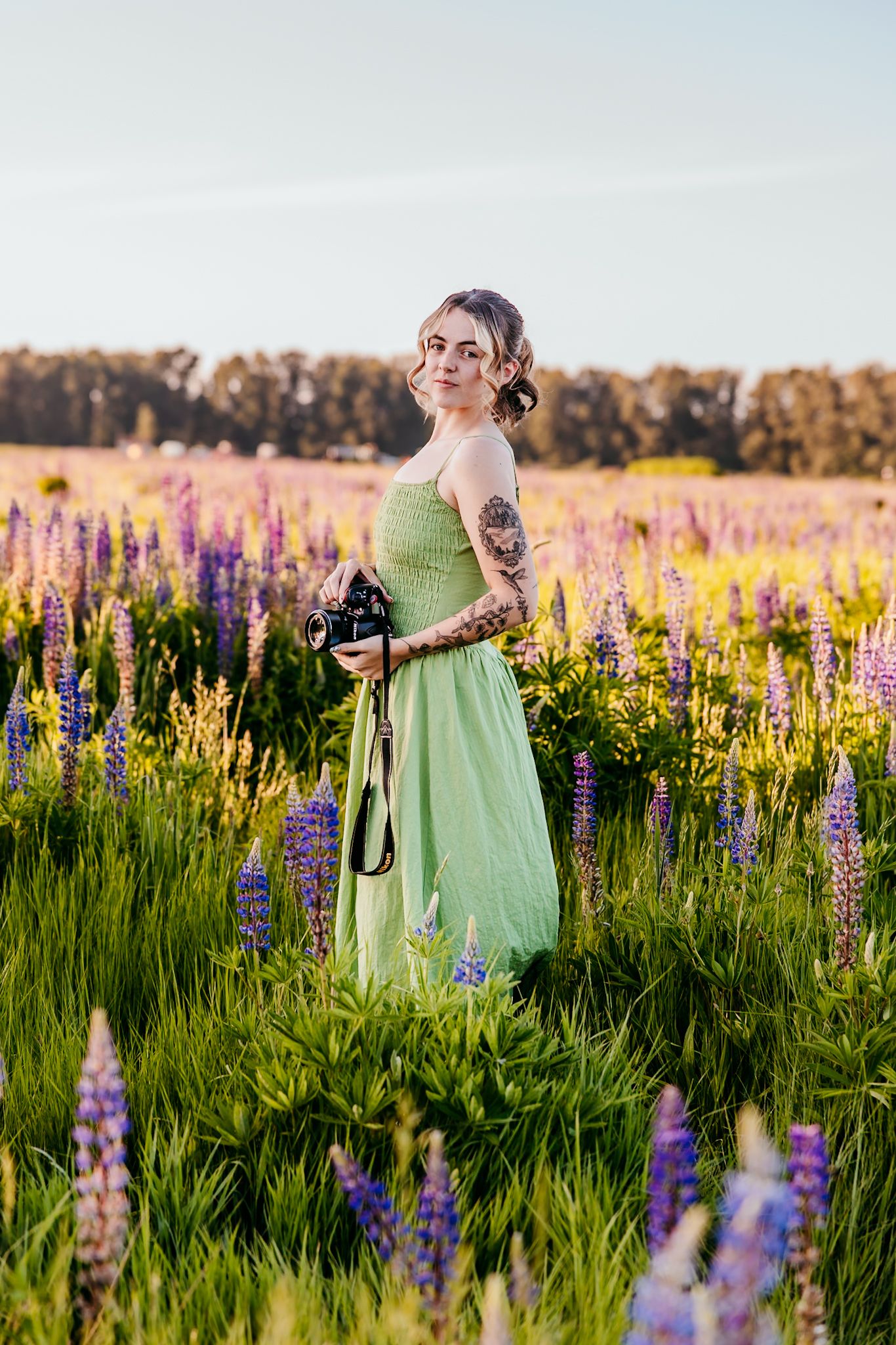 Tattooed woman in green dress holding camera stands in vibrant lupine flower field at golden hour.