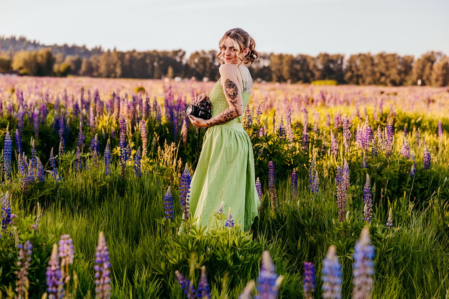 Woman in green dress standing in vibrant lupine flower field at golden hour, holding a bouquet.