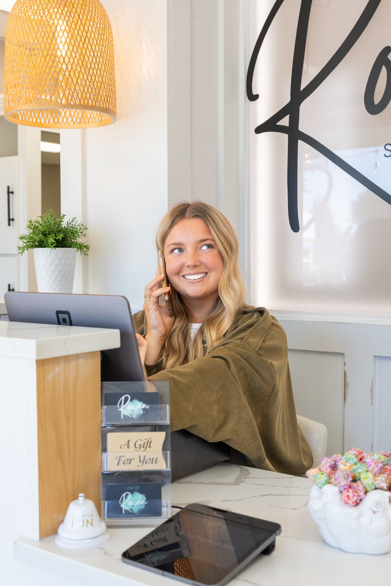 Smiling blonde woman talking on phone at a bright reception desk with gift boxes and a laptop.
