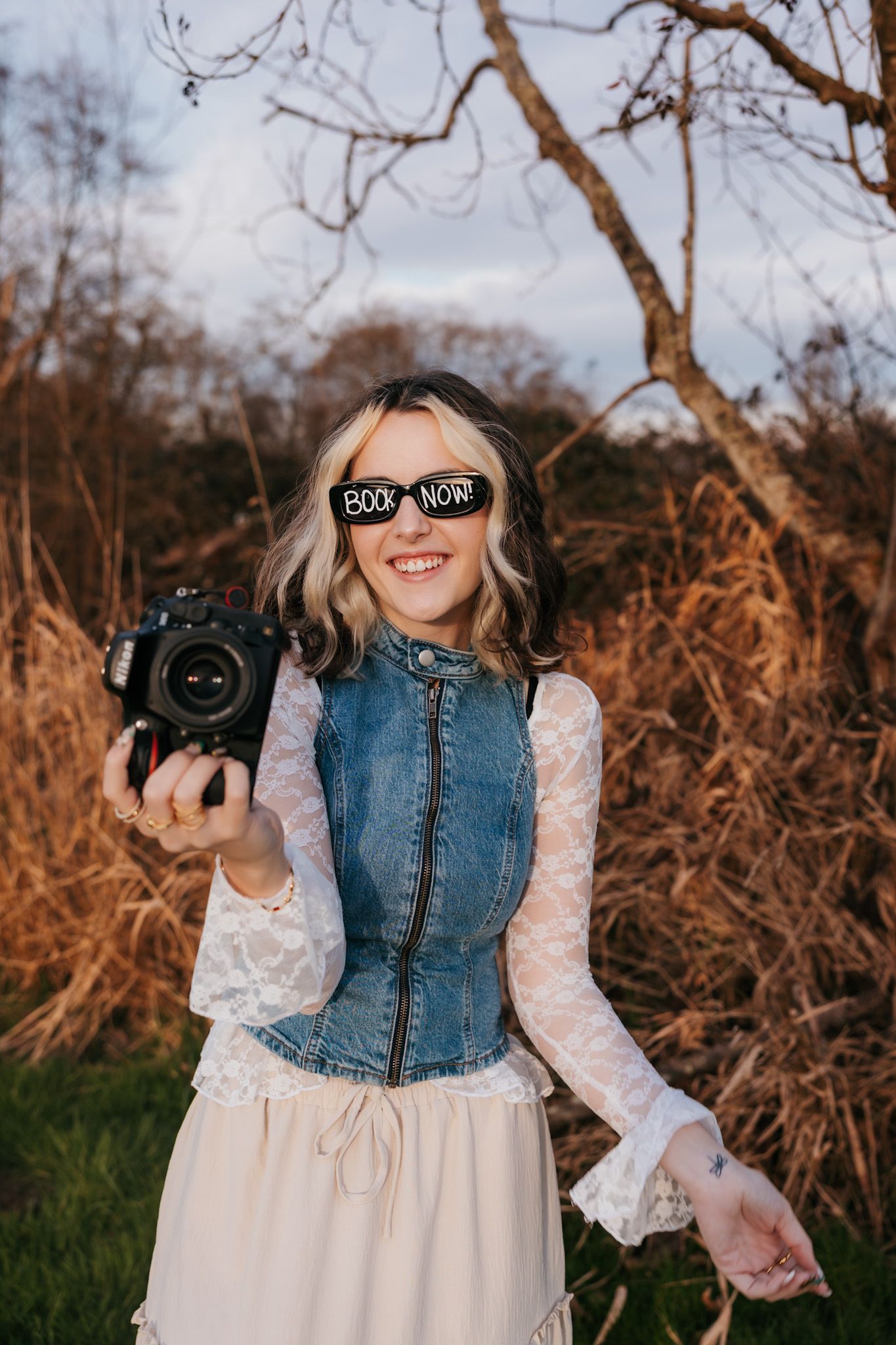 A photographer in a white lace dress and denim vest holds a camera during golden hour outdoor photoshoot.