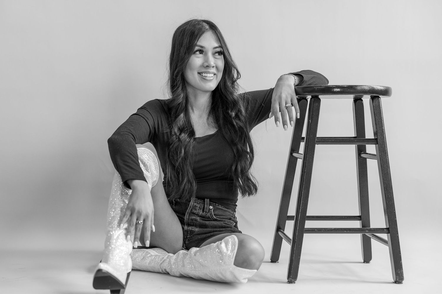 Black and white photo of a young woman with long hair sitting on floor beside a wooden stool, smiling confidently.