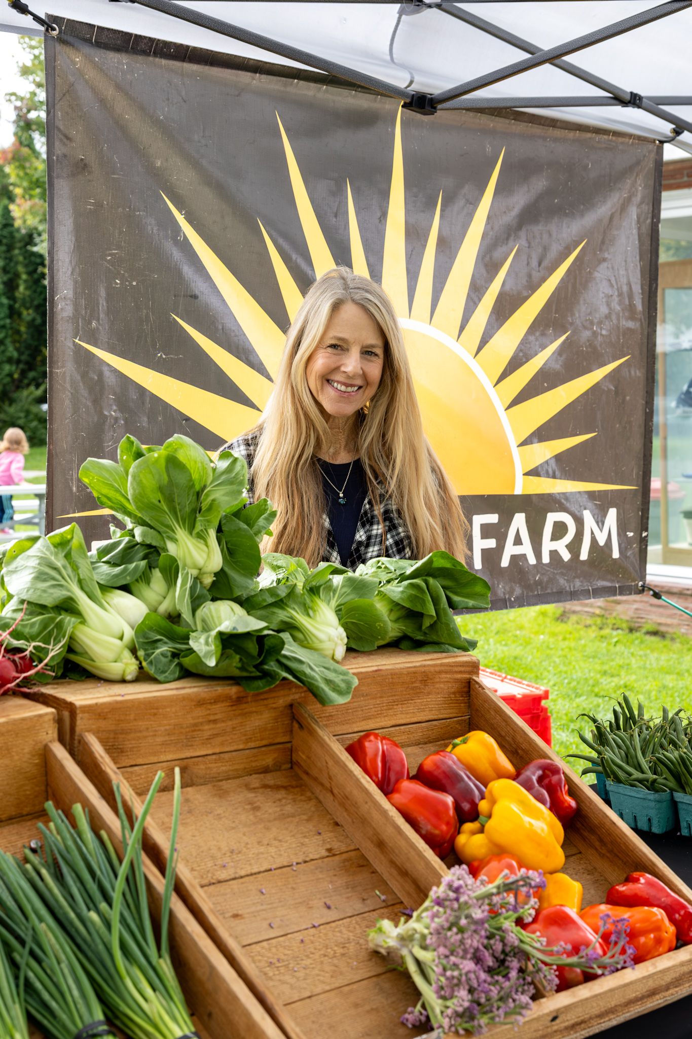Smiling woman at a farm market stand with fresh bok choy, bell peppers, and vegetables under a sun logo banner.